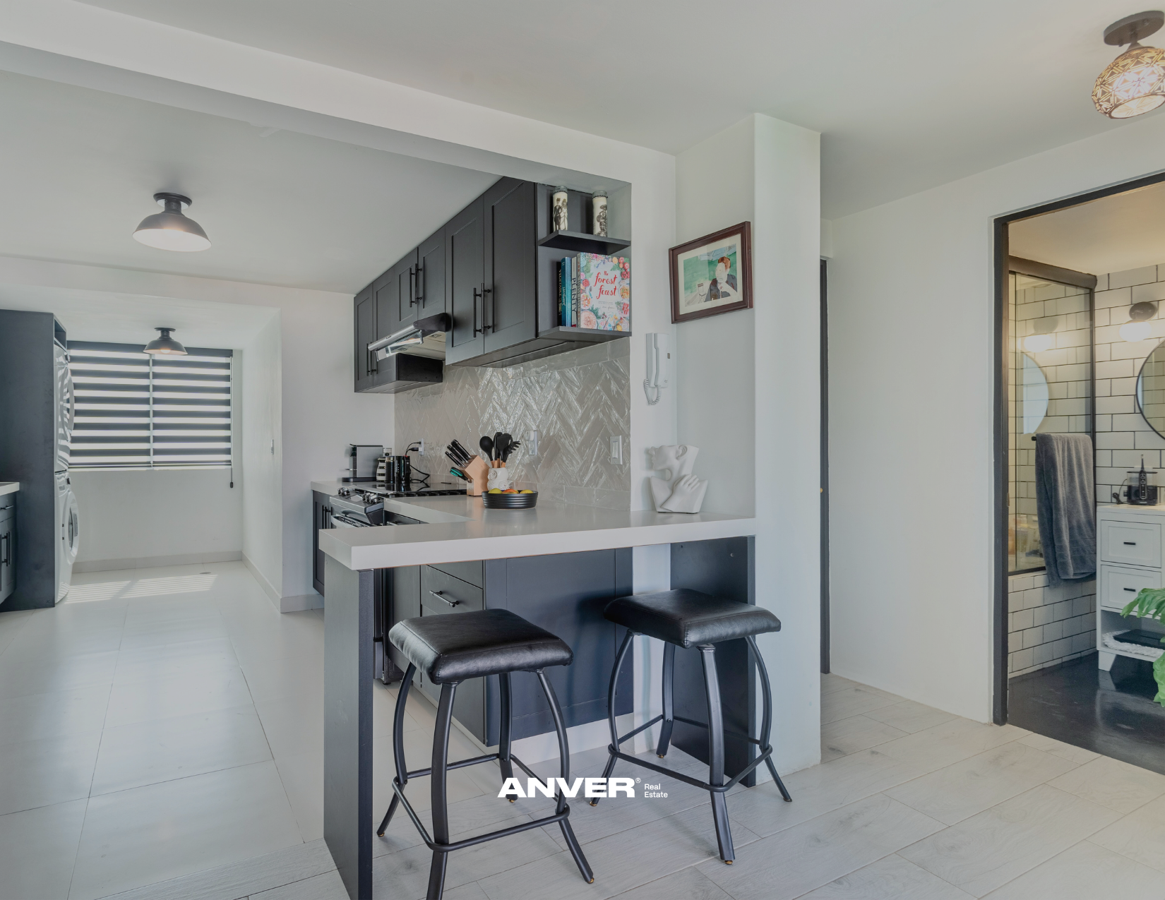 Cocina moderna con gabinetes negros, barra con tres taburetes, detalles decorativos y electrodomésticos de acero inoxidable, vista hacia otra habitación con baño en el fondo.