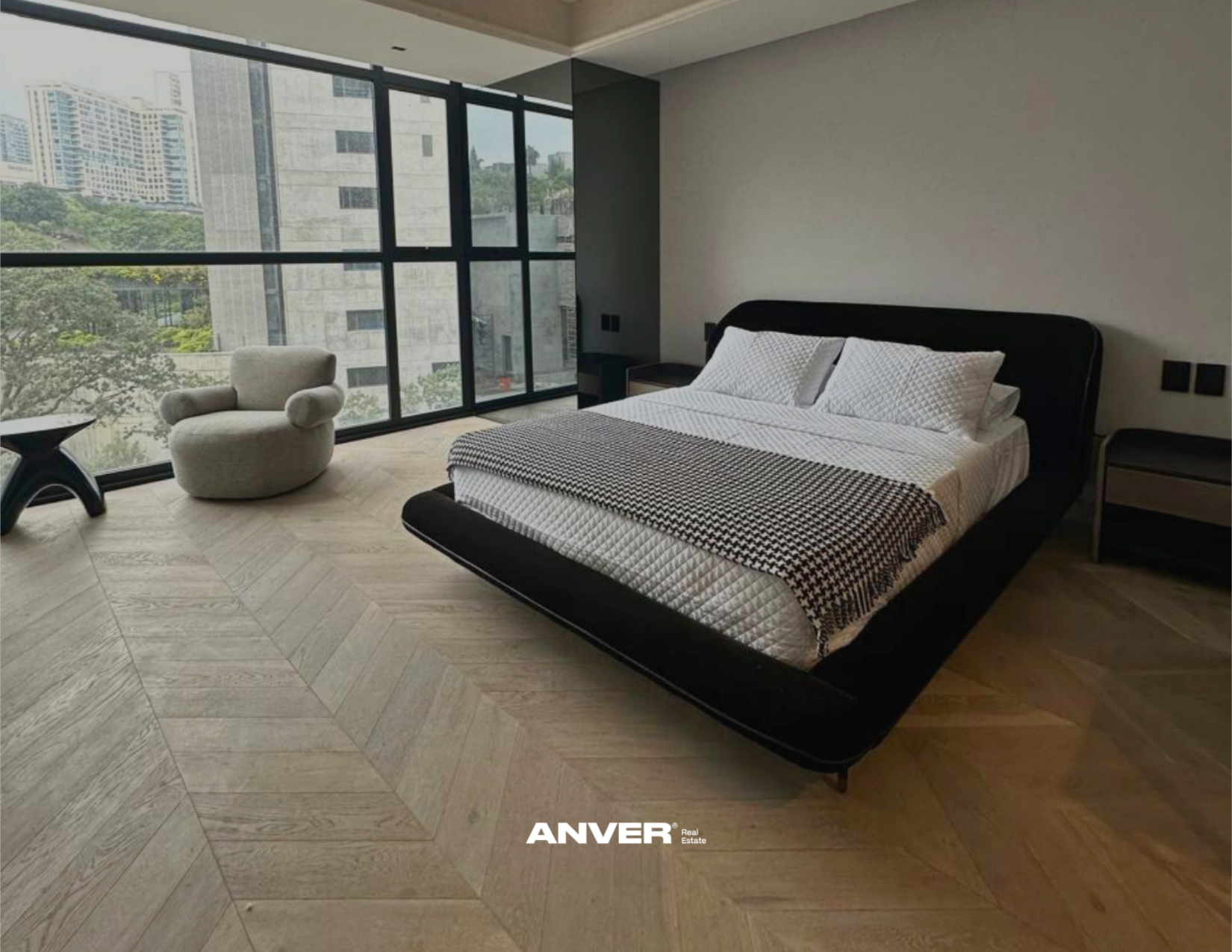 Habitación moderna con cama grande, cómodos almohadones, silla de estilo contemporáneo, vista de edificios y árboles a través de ventanas amplias, suelo de madera clara.