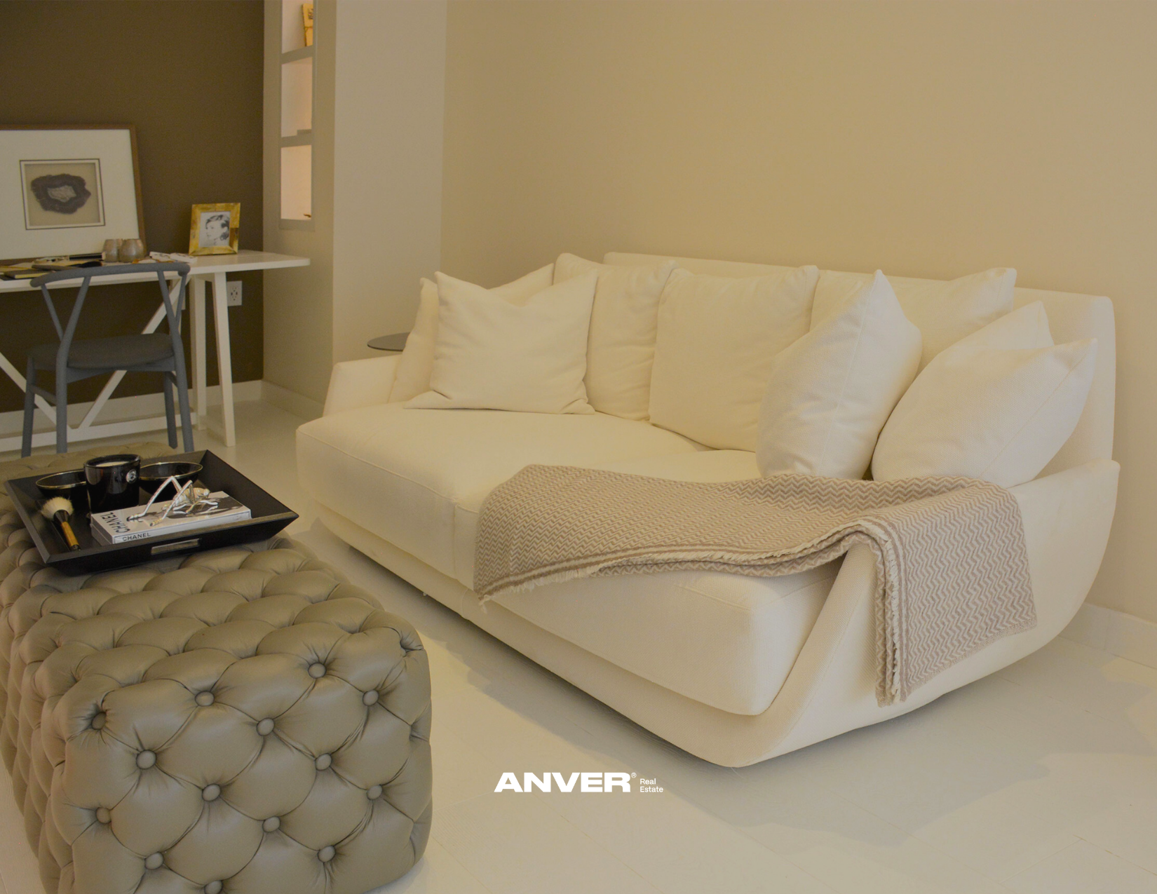 Sala de estar con sillón blanco, cojines blancos y una manta beig. Mesa con objetos y cuadros en la pared. Ottoman color beige. Logo de ANVER en la parte inferior.