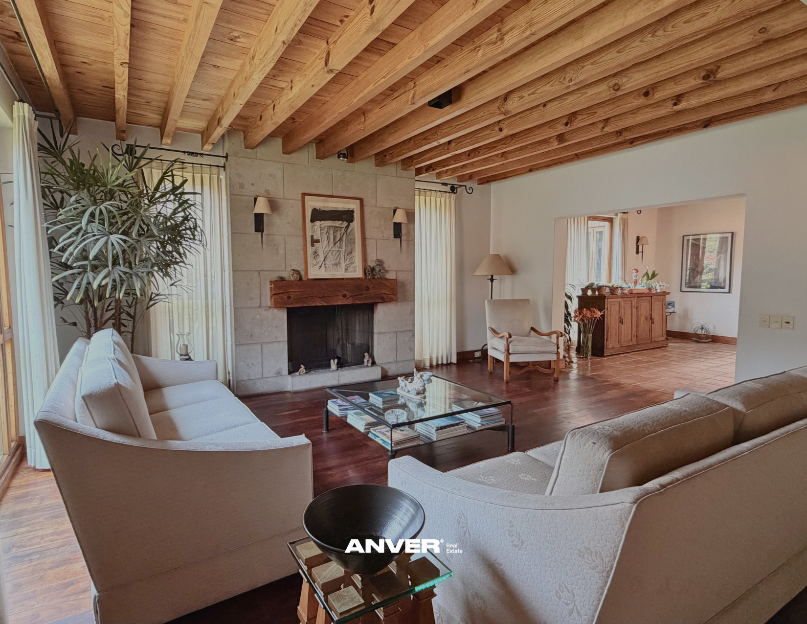 Sala de estar con paredes de piedra y madera en el techo, mobiliario beige y detalles decorativos, con ventanas con cortinas claras