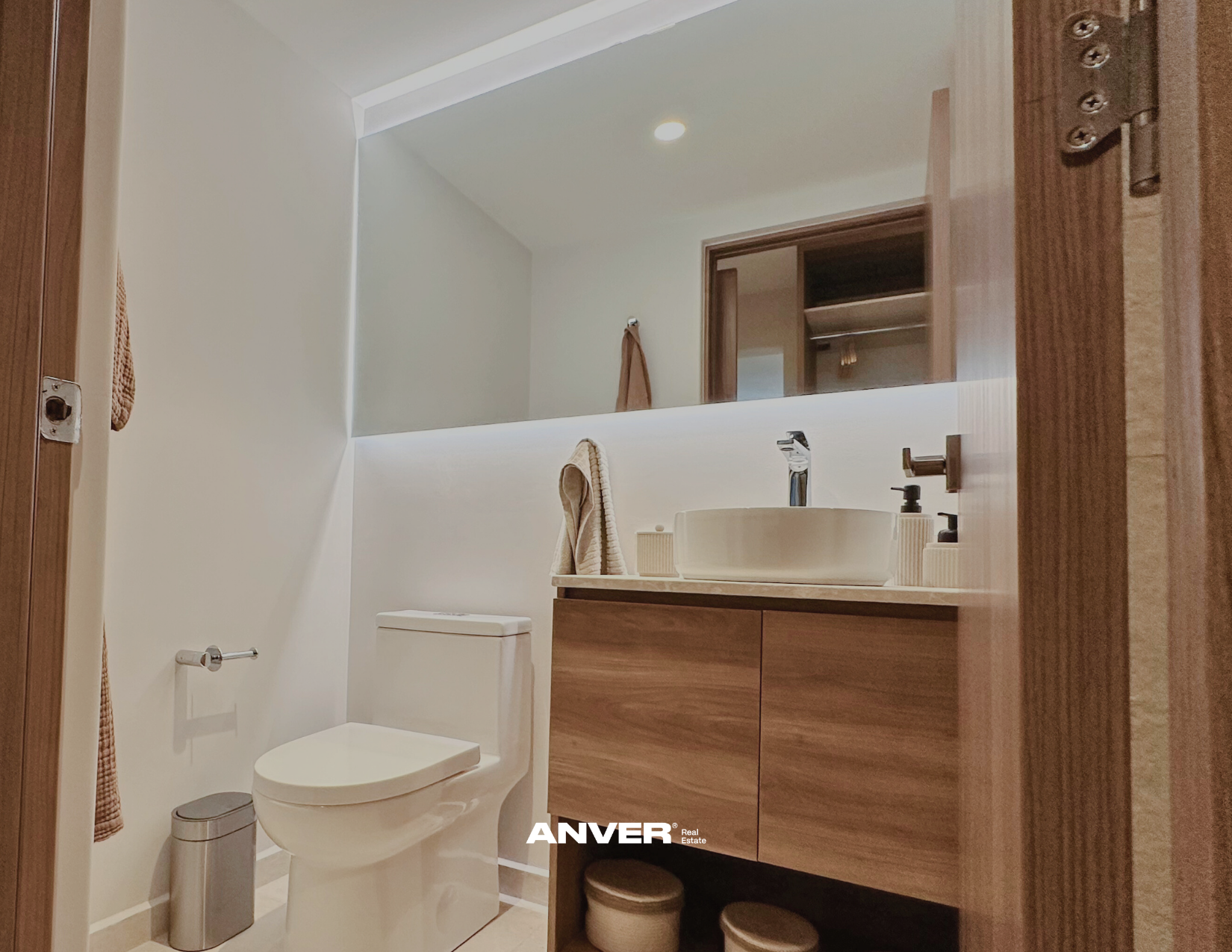 Baño con lavabo, espejo grande, inodoro, toallas, y gabinetes de madera, paredes blancas, iluminación moderna.
