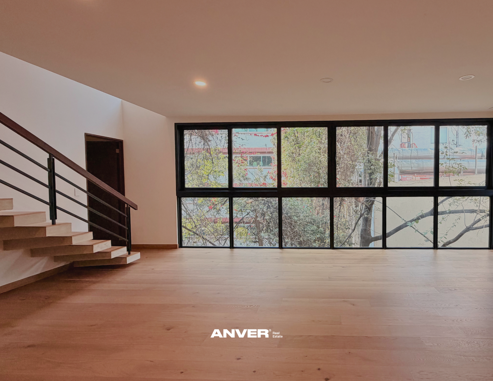 Interior moderno con pisos de madera, escalera de madera y barandilla negra, grandes ventanas de cristales que muestran árboles y edificios exteriores.