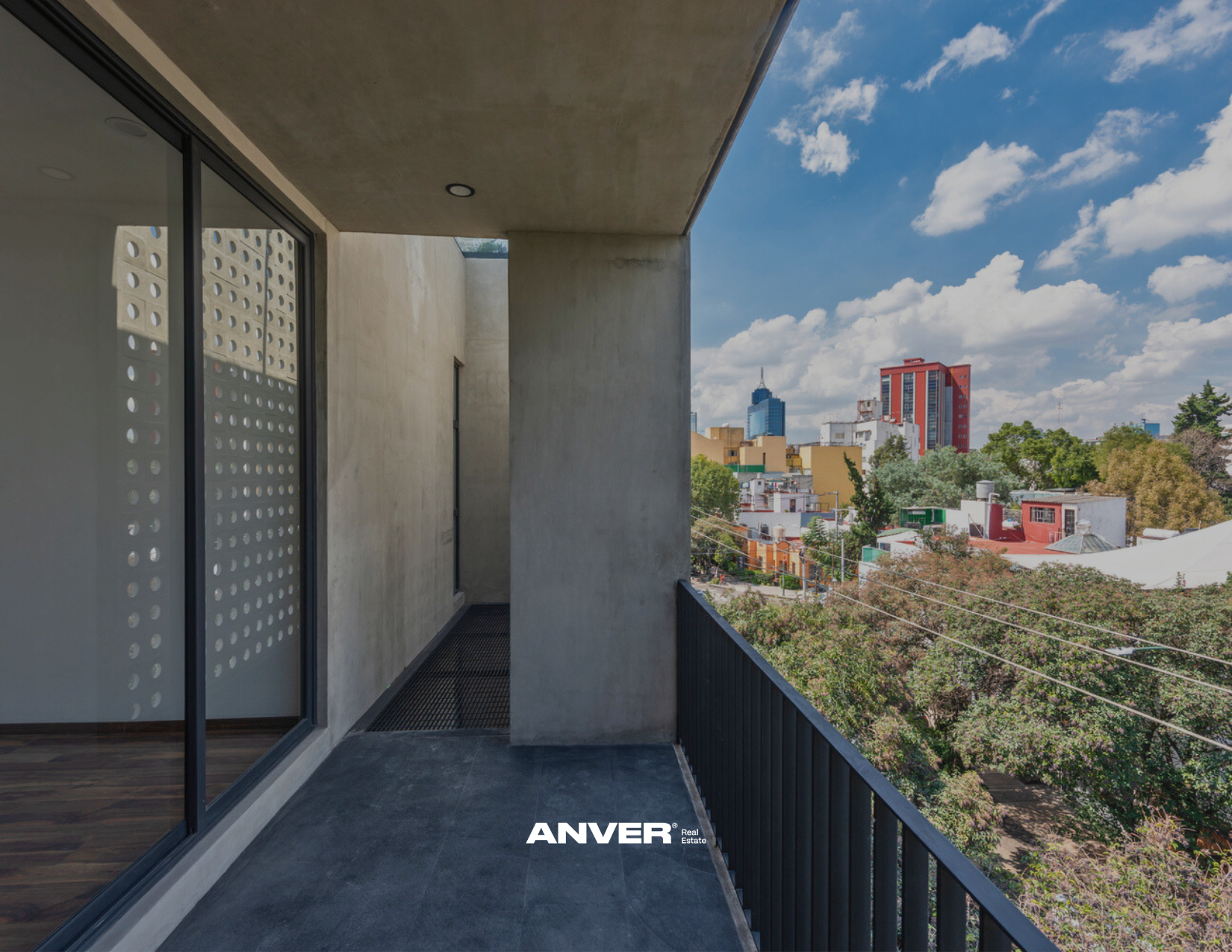 Balcon semi-cerrado en un apartamento con vista a la ciudad, árboles y edificios, cielo despejado con nubes.
