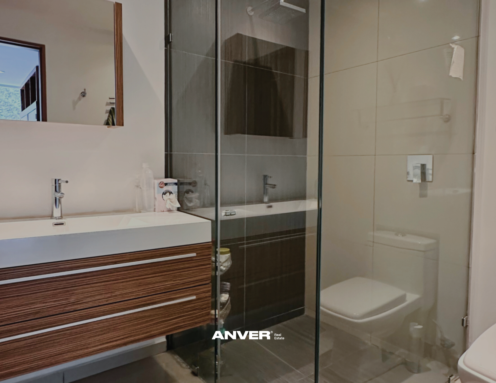 Baño moderno con lavabo de cerámica en madera, espejo, ducha con puertas de vidrio, inodoro y paredes de azulejo beige.