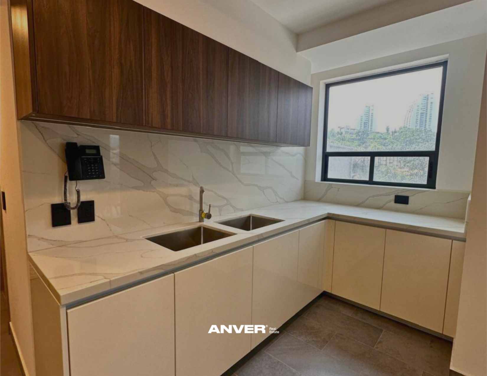 Cocina moderna con encimera de piedra con vetas, gabinetes de color beige y madera, doble fregadero y ventana grande que da vista a edificios altos y árboles.