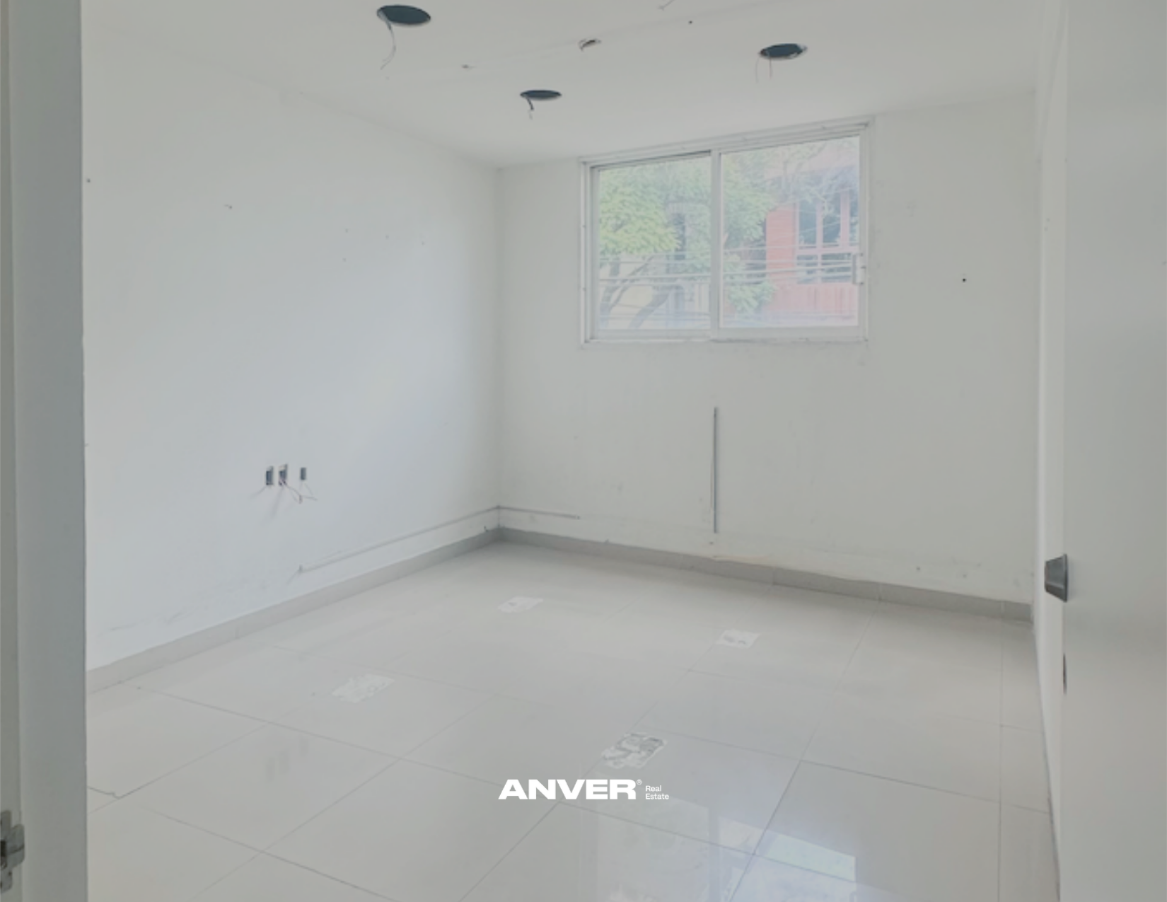 Interior de una habitación vacía en construcción con piso de cerámica blanca y paredes blancas, sin muebles ni decoración, con un ventanal grande que deja entrar luz natural.