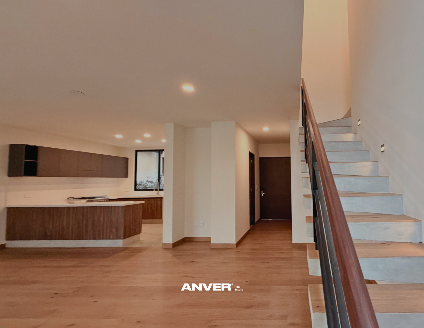 Interior de una casa con escalera de madera, pasillo y puerta oscura, con paredes blancas y suelo de madera.