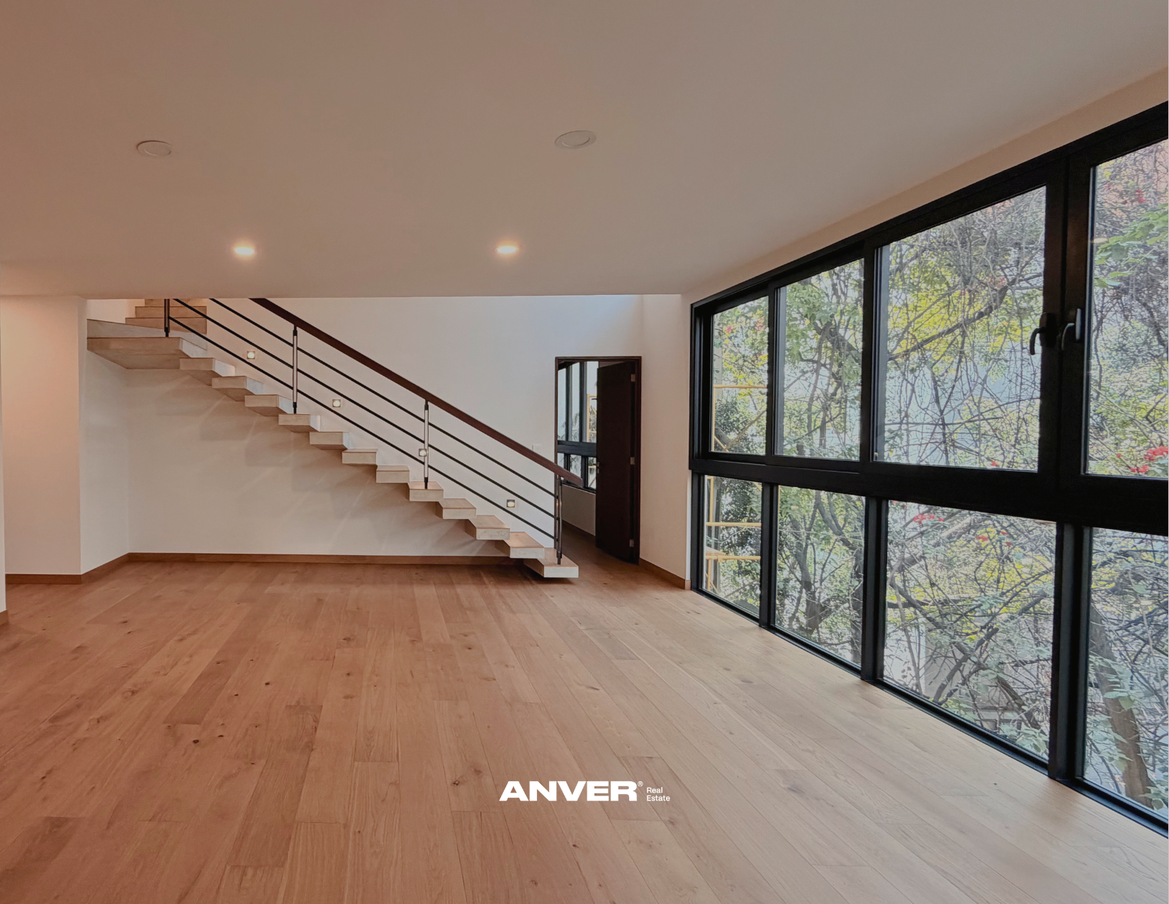 Interior de una casa moderna con pisos de madera y ventanas grandes que muestran árboles exteriores. Escalera con barandal de metal y madera. La pared y el techo son blancos, con luces empotradas. La imagen lleva un logotipo de 'ANVER' en la parte inferior.