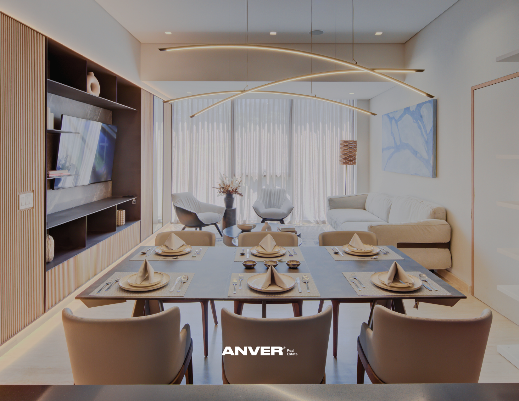 Sala de estar moderna con ventanas grandes con cortinas blancas, sillones blancos y mesa de centro con decoración, pared con cuadro azul y sofá beige, mesa de comedor con platos y cubiertos para seis personas, luz decorativa en el techo y estantería con objetos decorativos.