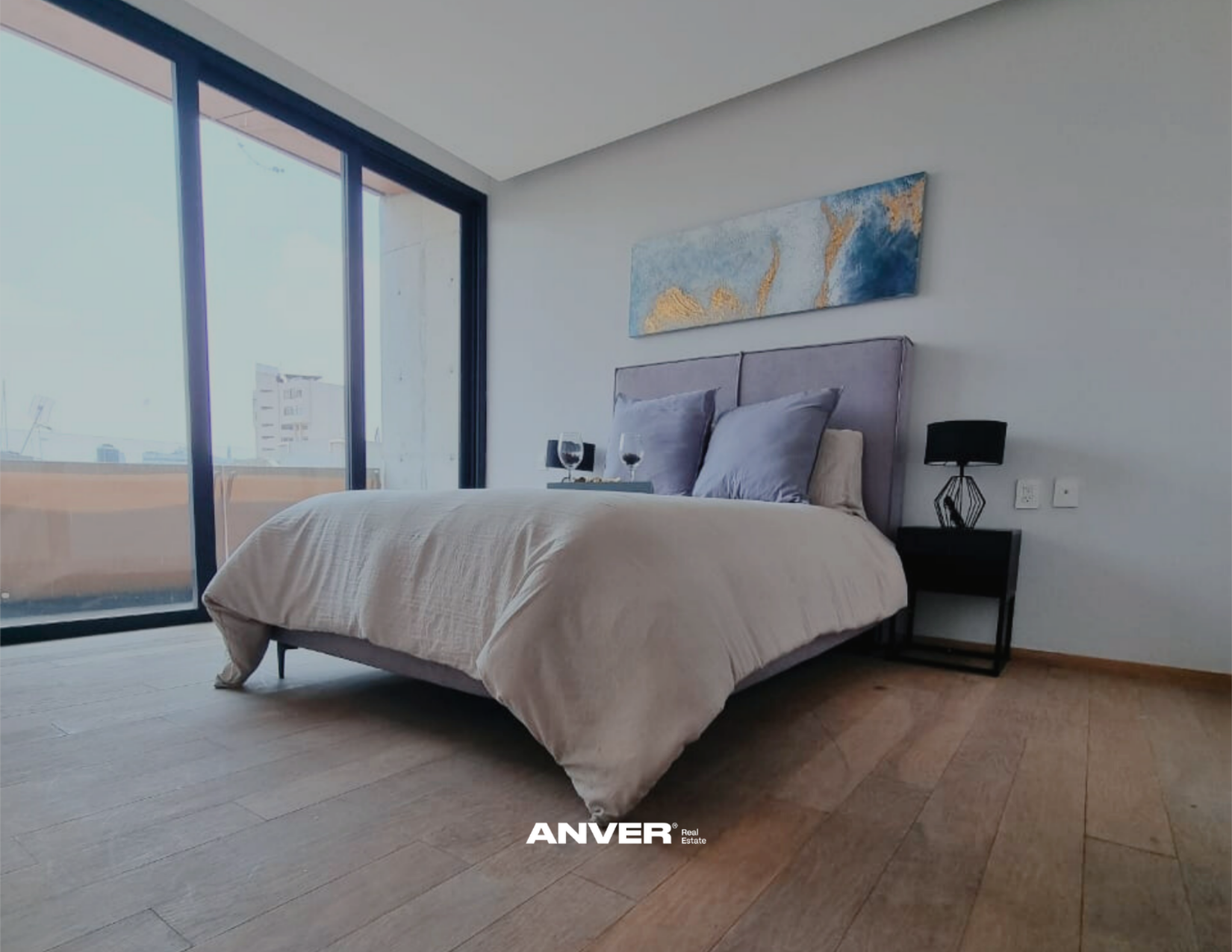 Habitación moderna con cama grande, cabecero acolchado, dos almohadas grises, dos copas de vino en una bandeja y ventana grande con vista a edificios.