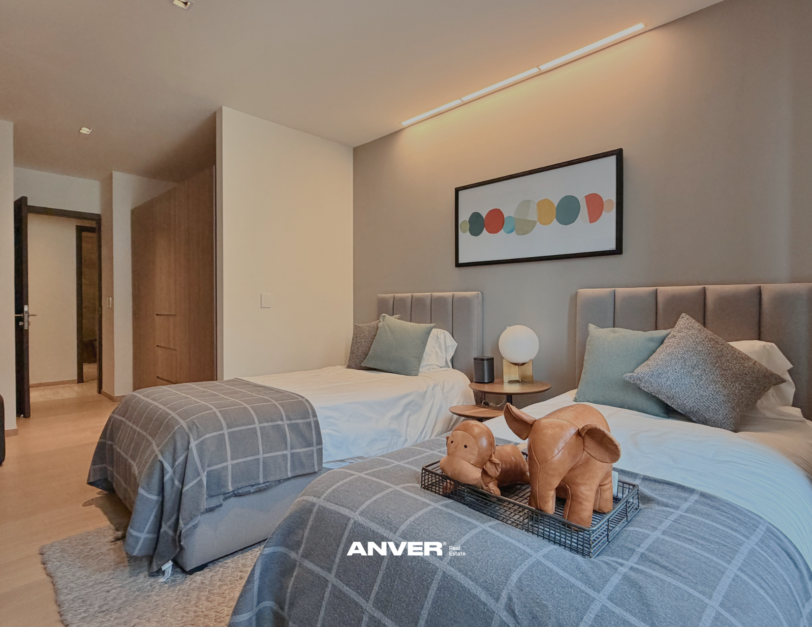 Habitación con dos camas con cabeceras tapizadas, ropa de cama gris con cuadros, almohadas grises y verdes, una lámpara de mesa entre las camas, cuadros abstractos en la pared, en la mesa junto a la cama hay una figura de perro de peluche y un cesto con artículos decorativos, piso de madera y una alfombra beige.