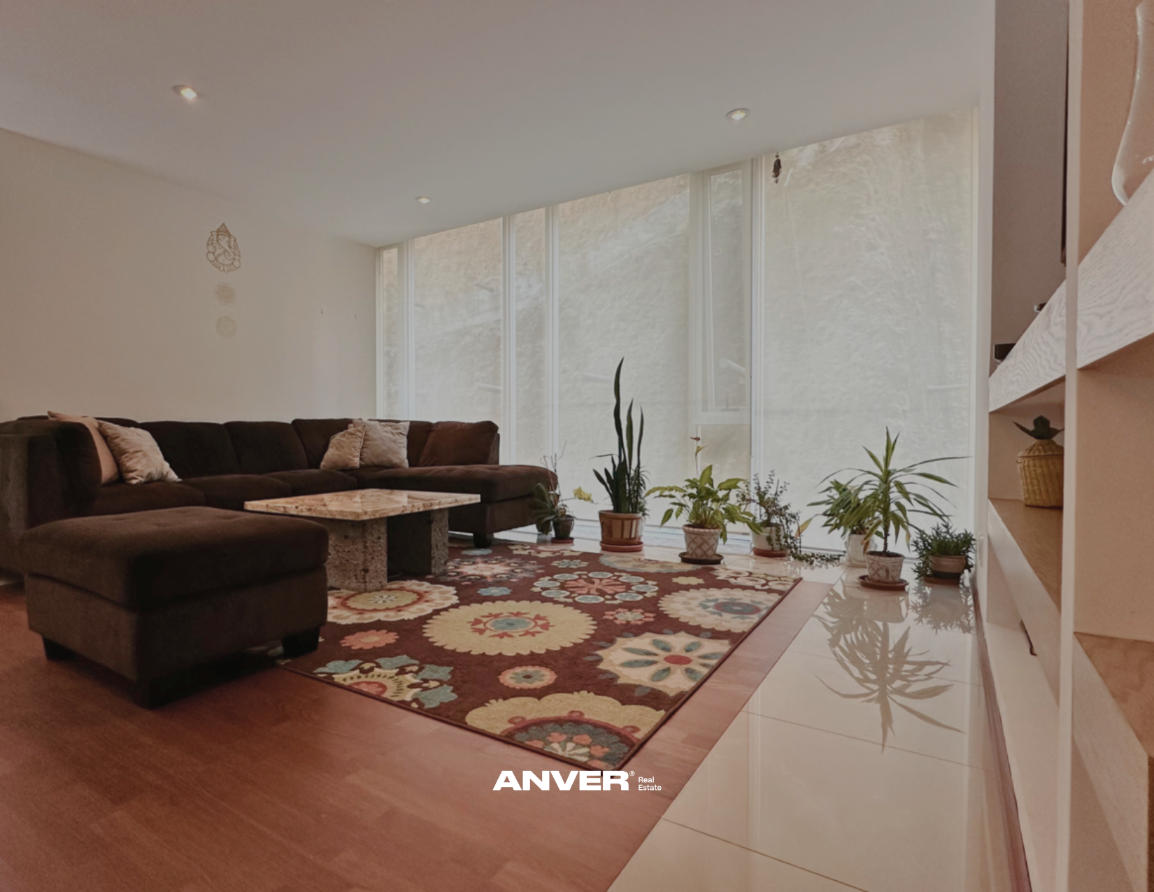 Sala de estar con sillón en forma de L, mesa de centro de piedra, alfombra con patrones coloridos, varias plantas en macetas, paredes blancas y ventanas con cortinas translúcidas, ambiente acogedor y luminoso