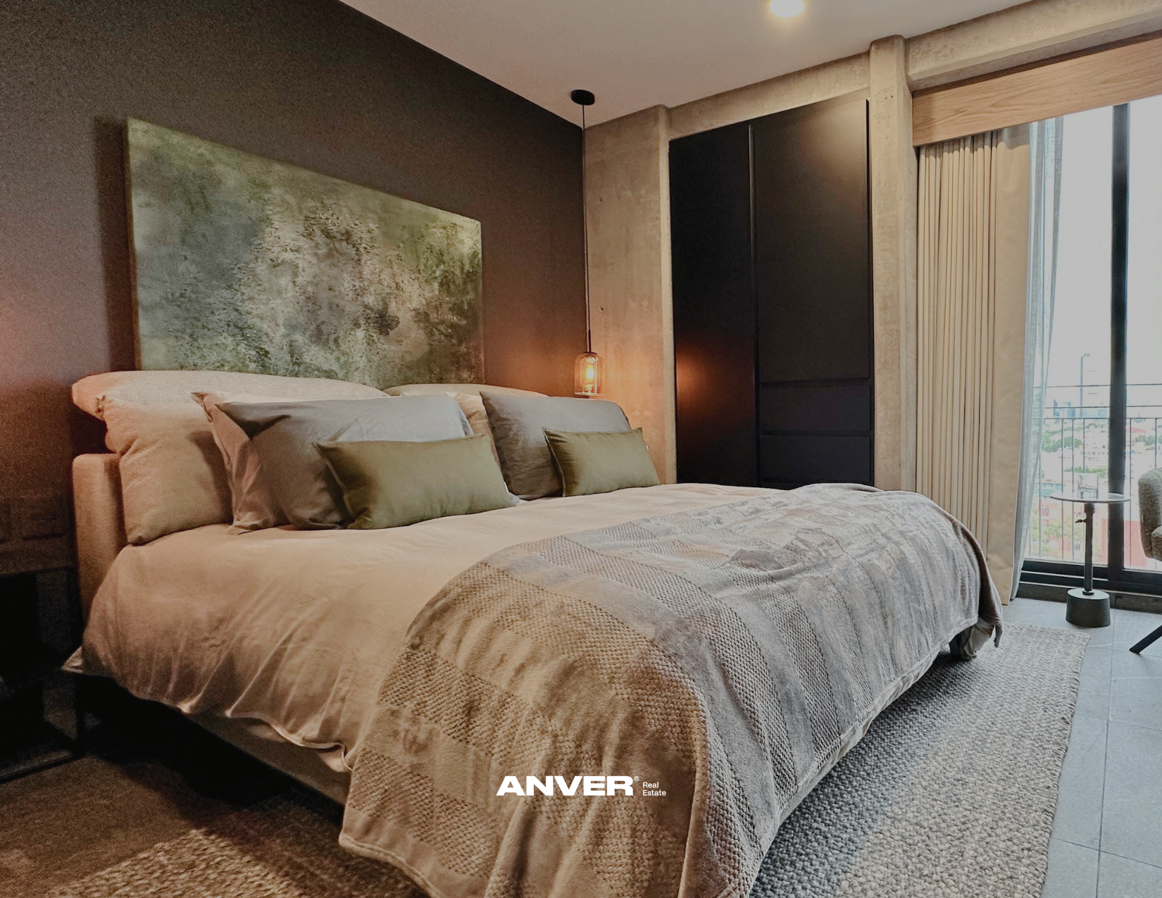 Habitación moderna con cama grande, almohadas verdes y grises, cabecera artística, lámpara colgante y ventana con vista a la ciudad