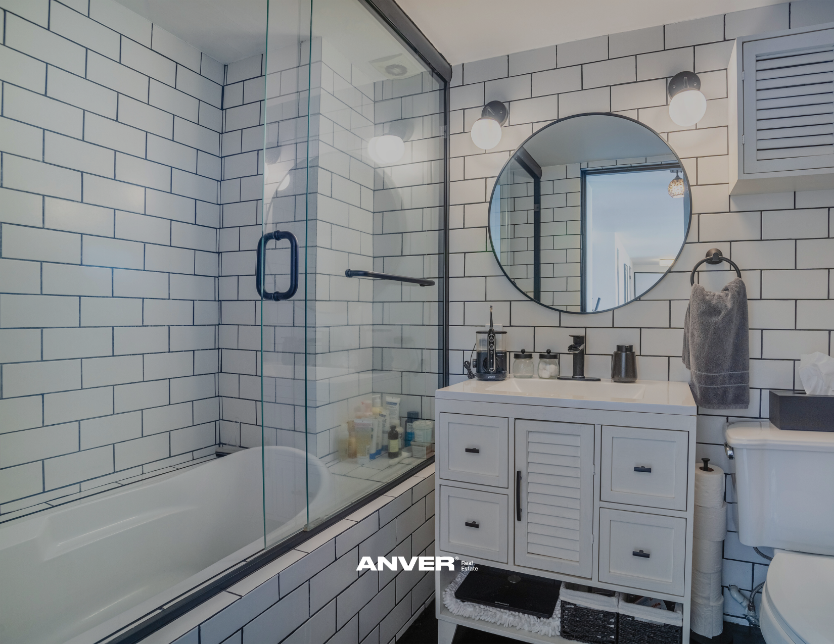 Baño con paredes de azulejos blancos con línea negra, ducha con puerta de cristal, lavabo con espejo redondo, varias luces, toalla gris en soporte negro, gabinetes blancos, y accesorios en negro.
