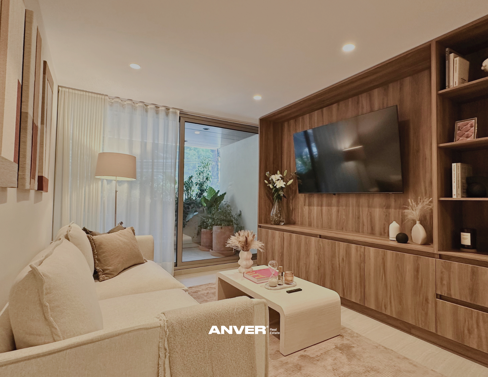 Sala de estar con sofá beige, mesa de centro blanca y decoración con flores y libros, pared de madera con televisión y estanterías, cortinas blancas, jardín visible desde el balcón.