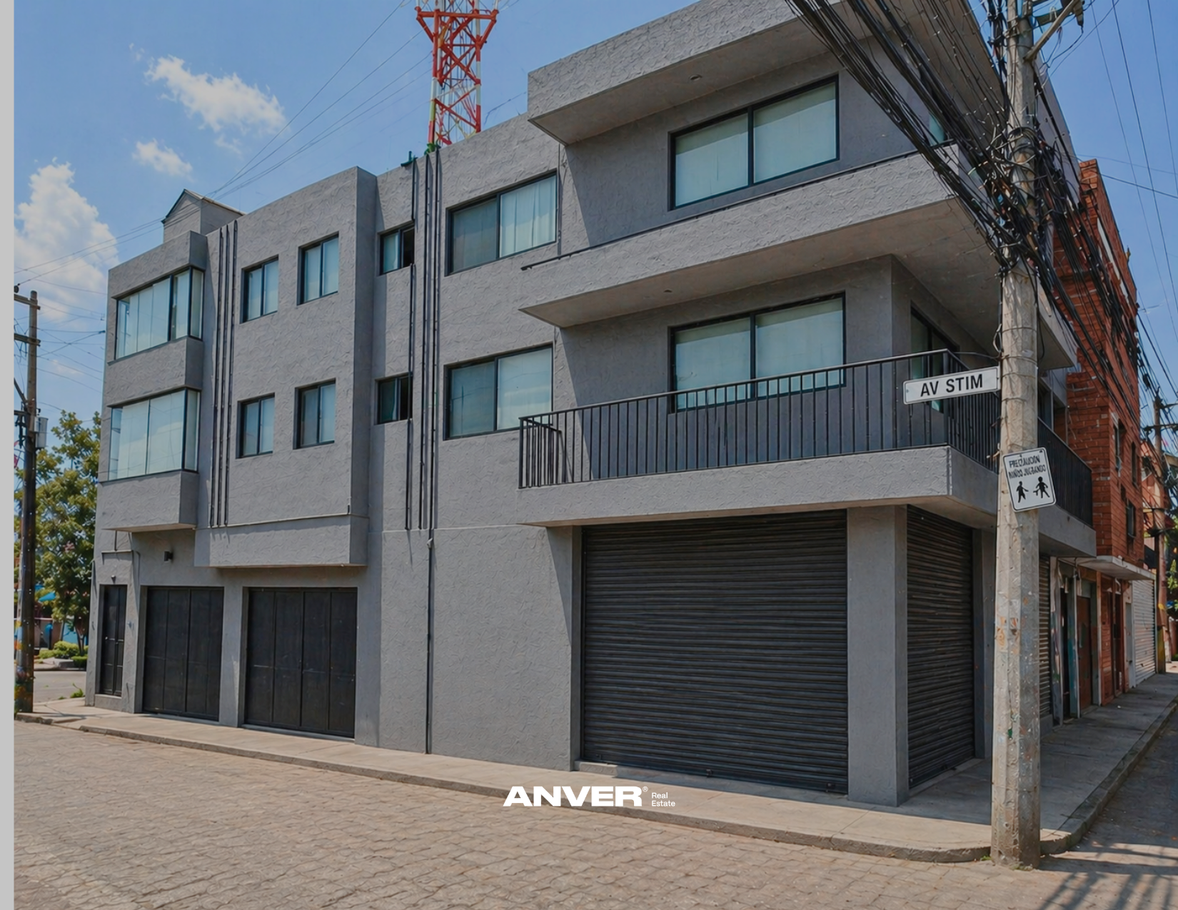 Edificio moderno de varios pisos con ventanas grandes y persianas negras, en una esquina de la calle, con anuncios de semáforo y de paso peatonal. Encima del edificio hay un poste de electricidad y una torre de comunicación. El cielo está despejado con algunas nubes.