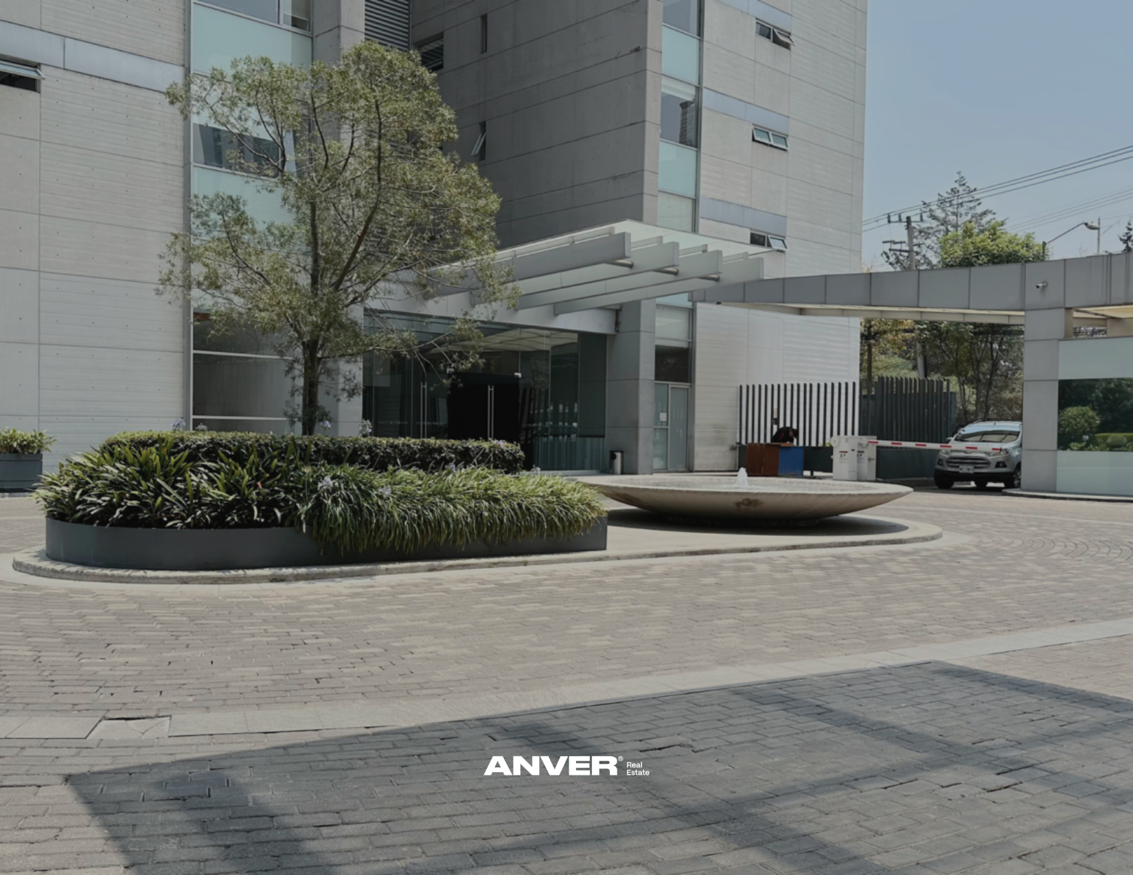 Vista exterior de un edificio moderno con área de entrada, áreas verdes y un vehículo estacionado, con señal de marca 'ANVER'. 