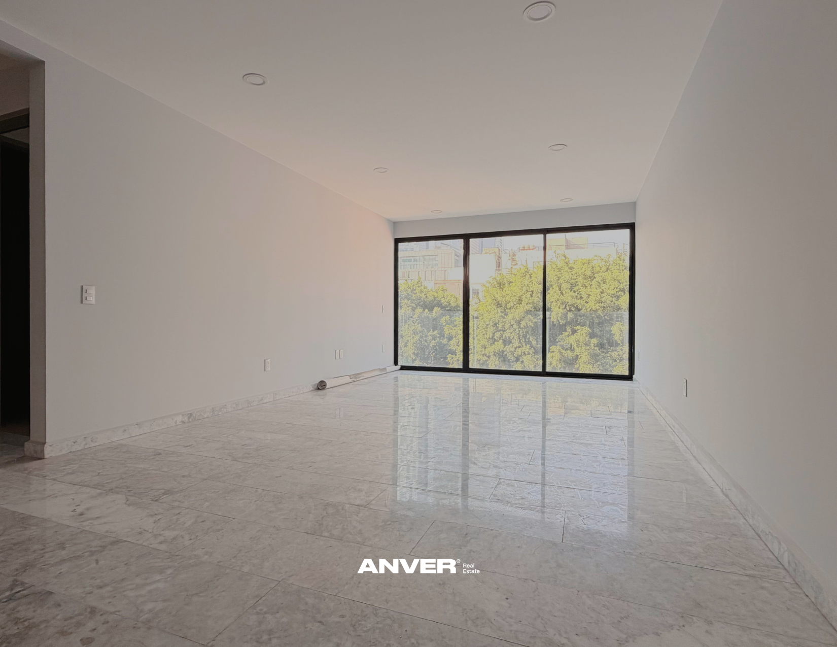 Sala vacía con paredes blancas, pisos de mármol y ventana grande con vista a árboles y edificios