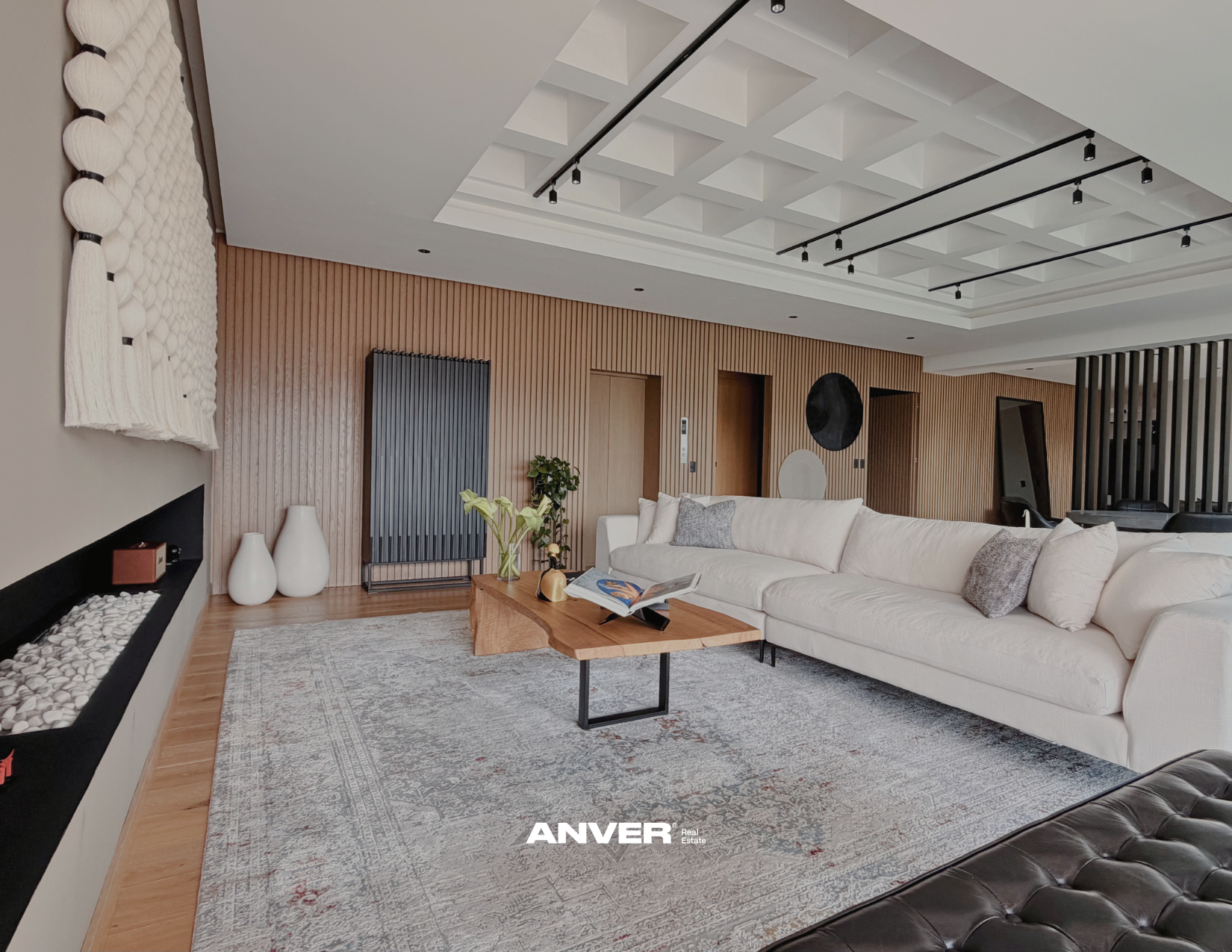 Sala de estar con sillón blanco, mesa de madera con libro y plantas, paredes de madera, decoración moderna y luminarias en el techo.