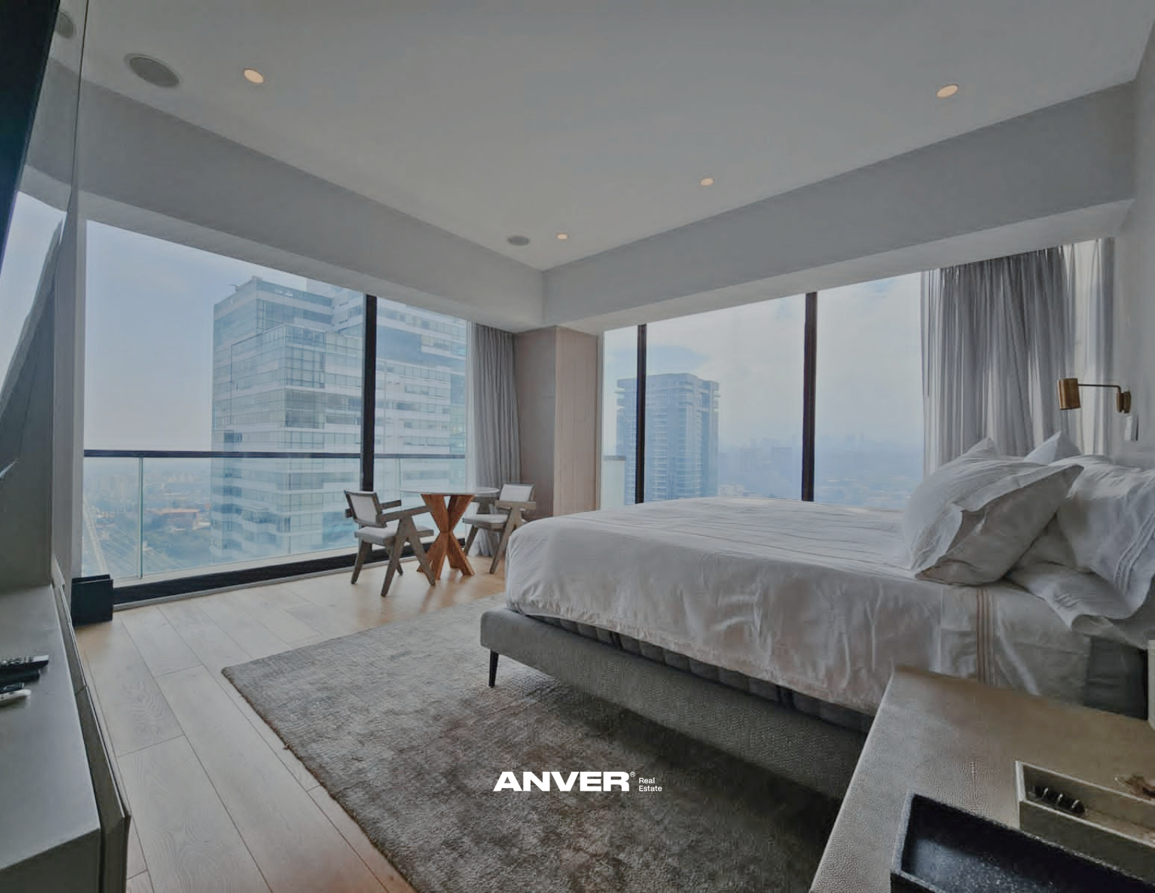 Habitación moderna con cama grande y ropa de cama blanca, mesas y sillas junto a ventanas grandes con vista a edificios altos en la ciudad.