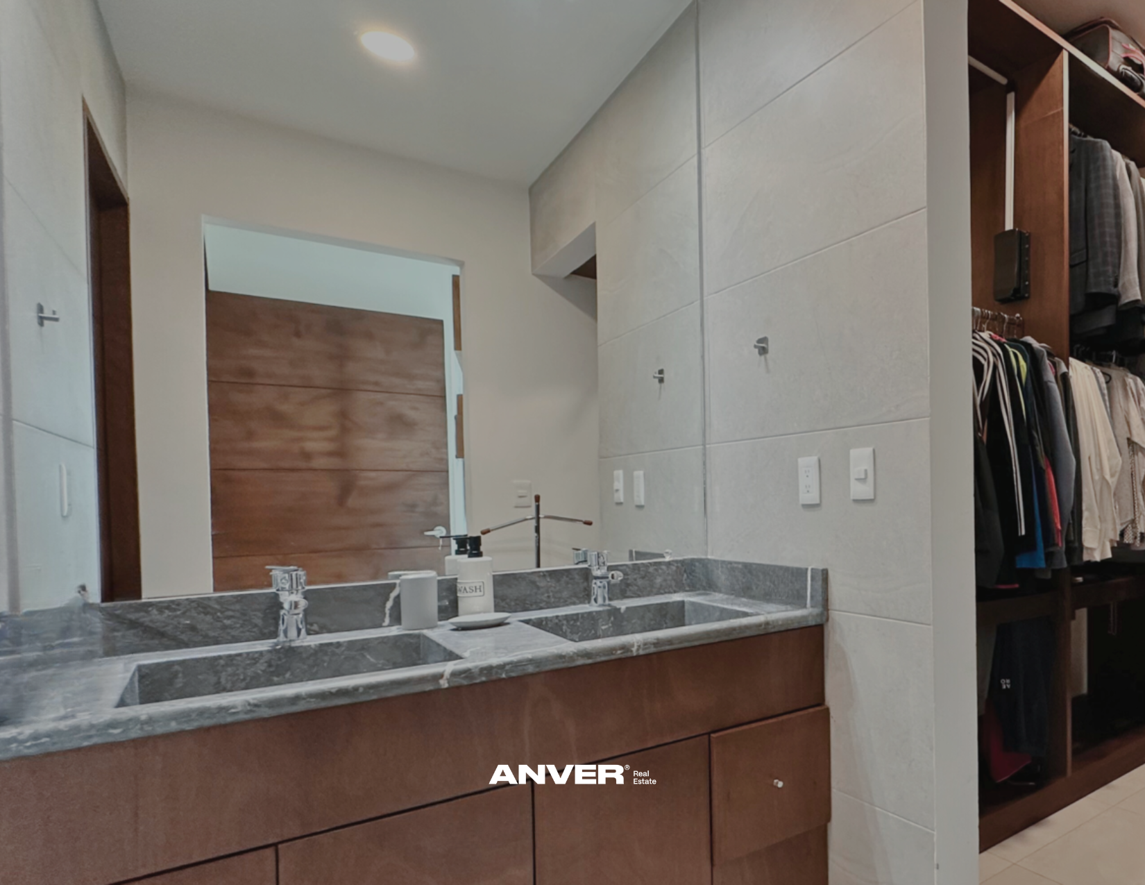 Imagen de un baño con lavabo de granito gris, espejos y paredes revestidas en azulejo beige, puerta de madera y closet con ropa en el fondo.