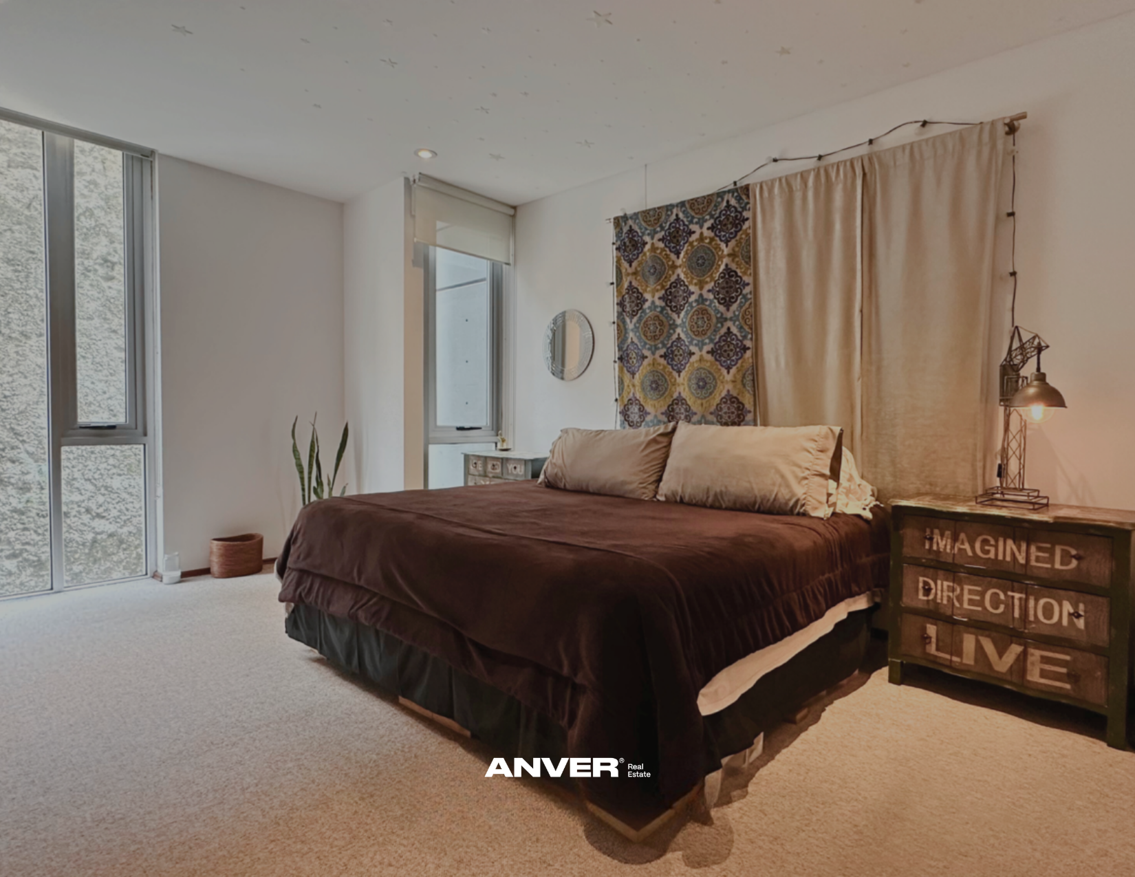Habitación con cama de tamaño king, dos almohadas grandes, cortinas beige y decorativas, lámpara de mesa con luz cálida, pared decorada con colcha y espejo redondo, planta en maceta, ventana grande con vistas a árboles, pared blanca y alfombra beige.