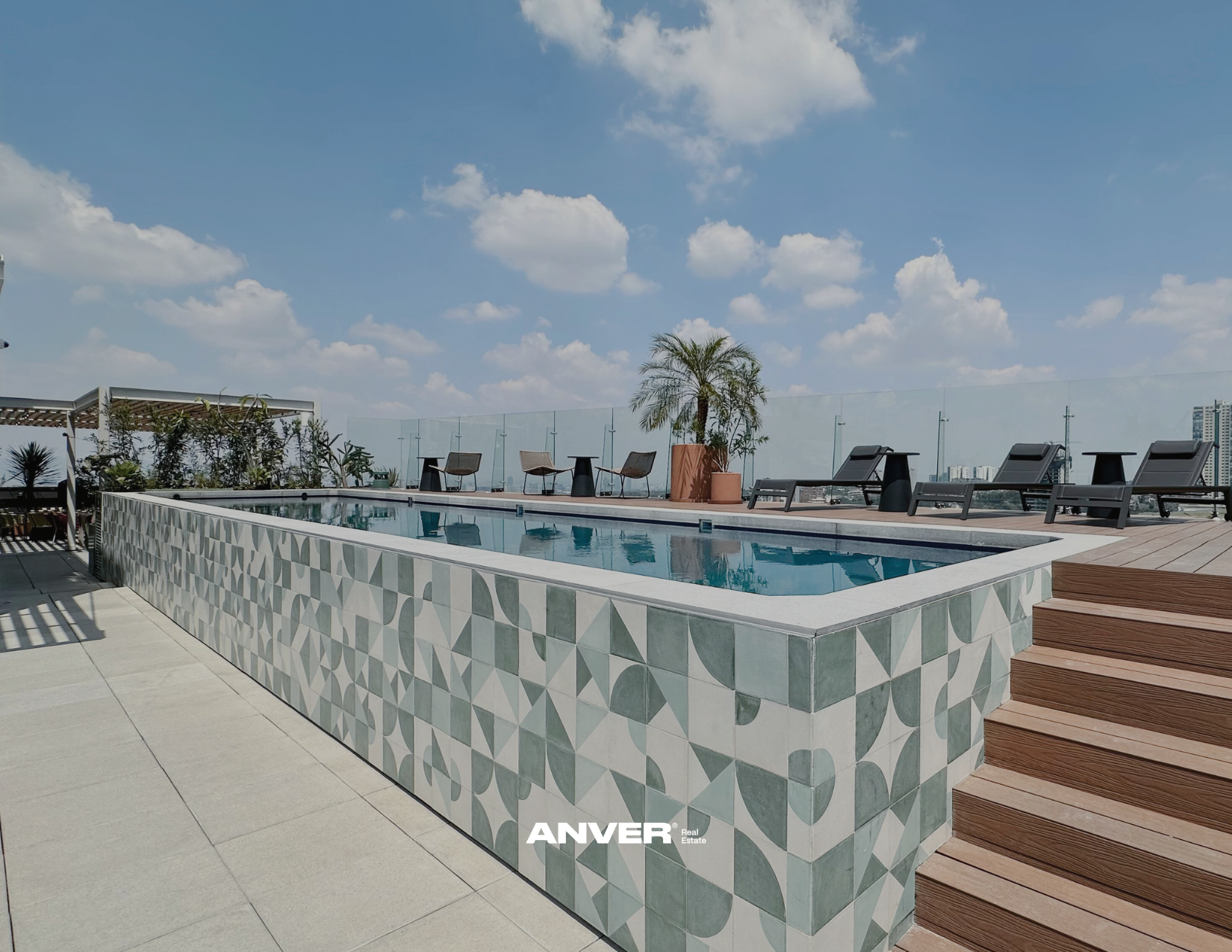 Piscina en la azotea con sillas y plantas, vista al cielo con nubes y ciudad al fondo.