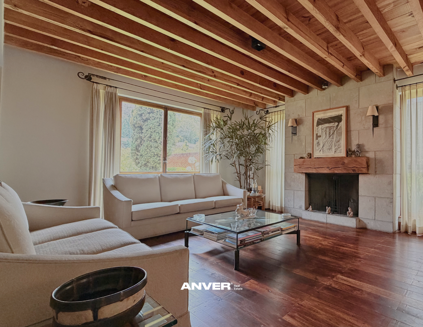 Sala de estar con sillones beige, mesa de centro de vidrio con libros, chimenea de piedra con un maderón arriba, una planta grande junto a la ventana y paredes blancas con cortinas claras, techo de madera con vigas expuestas.