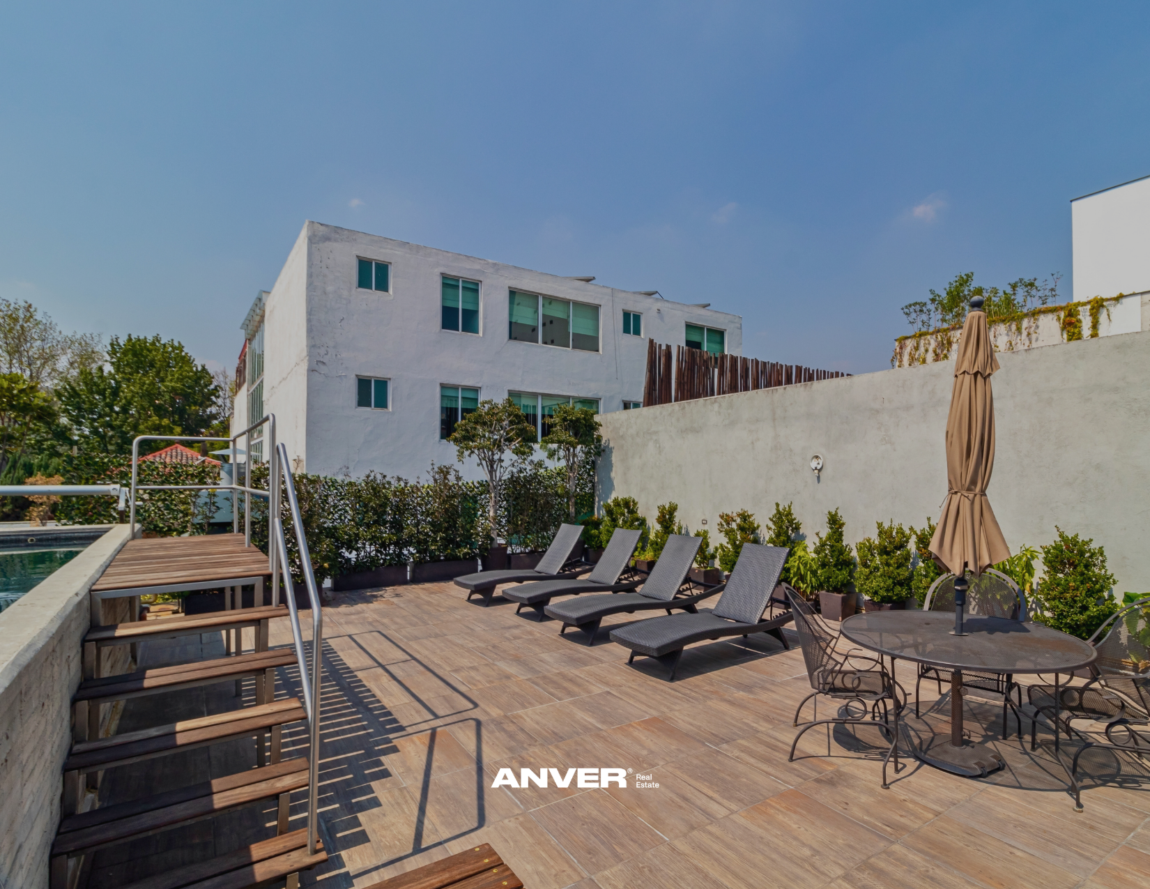 Área de alberca y jardín en una azotea, con sillones reclinables, mesa con sombrilla cerrada, plantas y edificio moderno al fondo, cielo despejado.