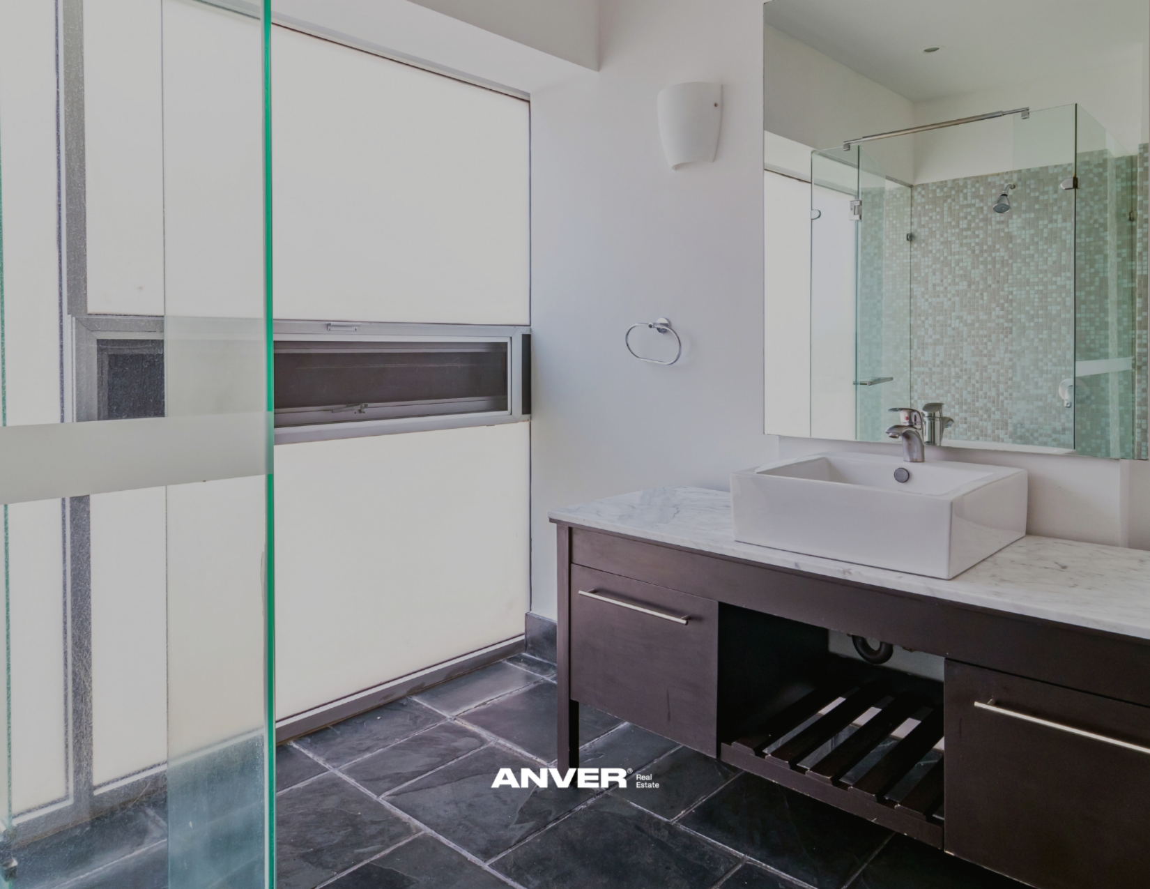 Baño con lavabo rectangular de cerámica en un mueble de madera, espejo grande, ducha con puertas de cristal y azulejos en tonos verdes y beige, pared blanca y piso de piedra oscura.