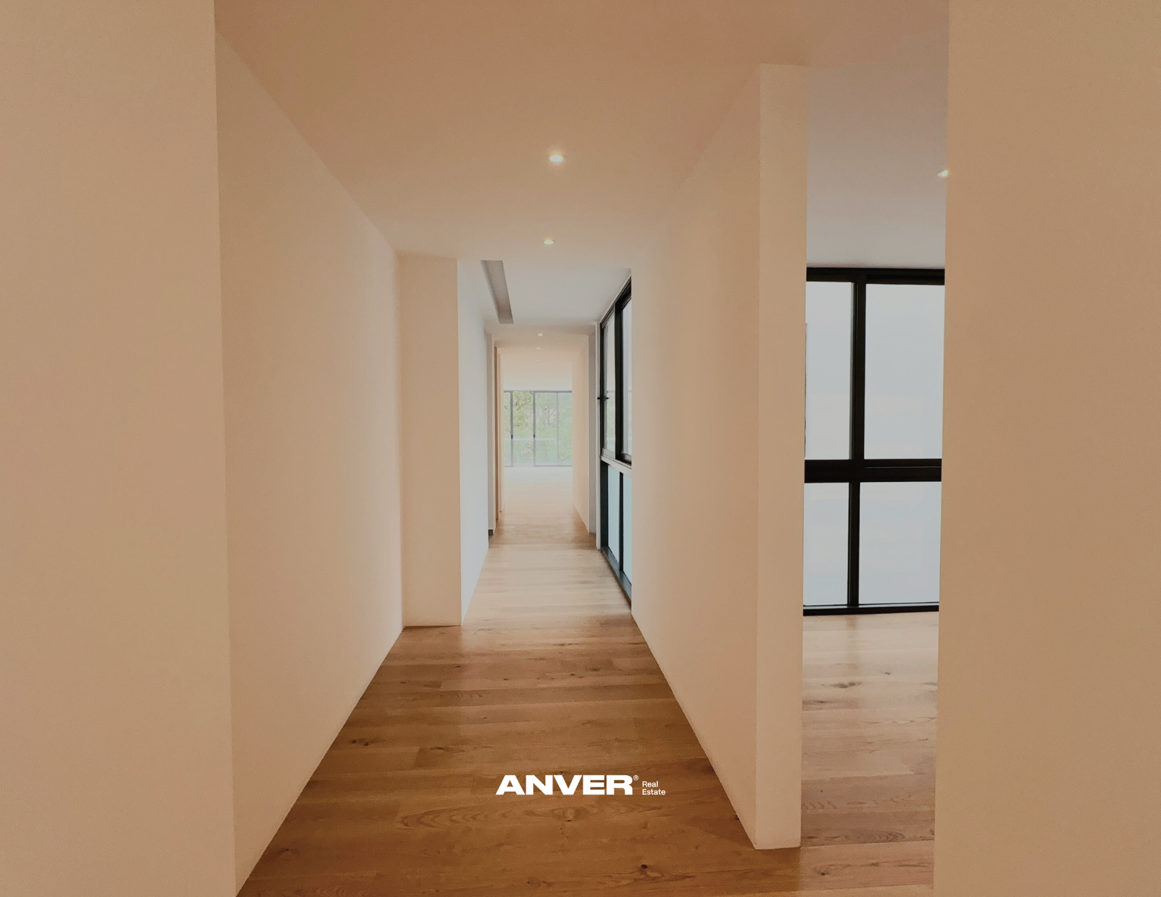 Pasillo interior con madera clara y paredes blancas, ventanas negras con vista al exterior y puertas corredizas de cristal.