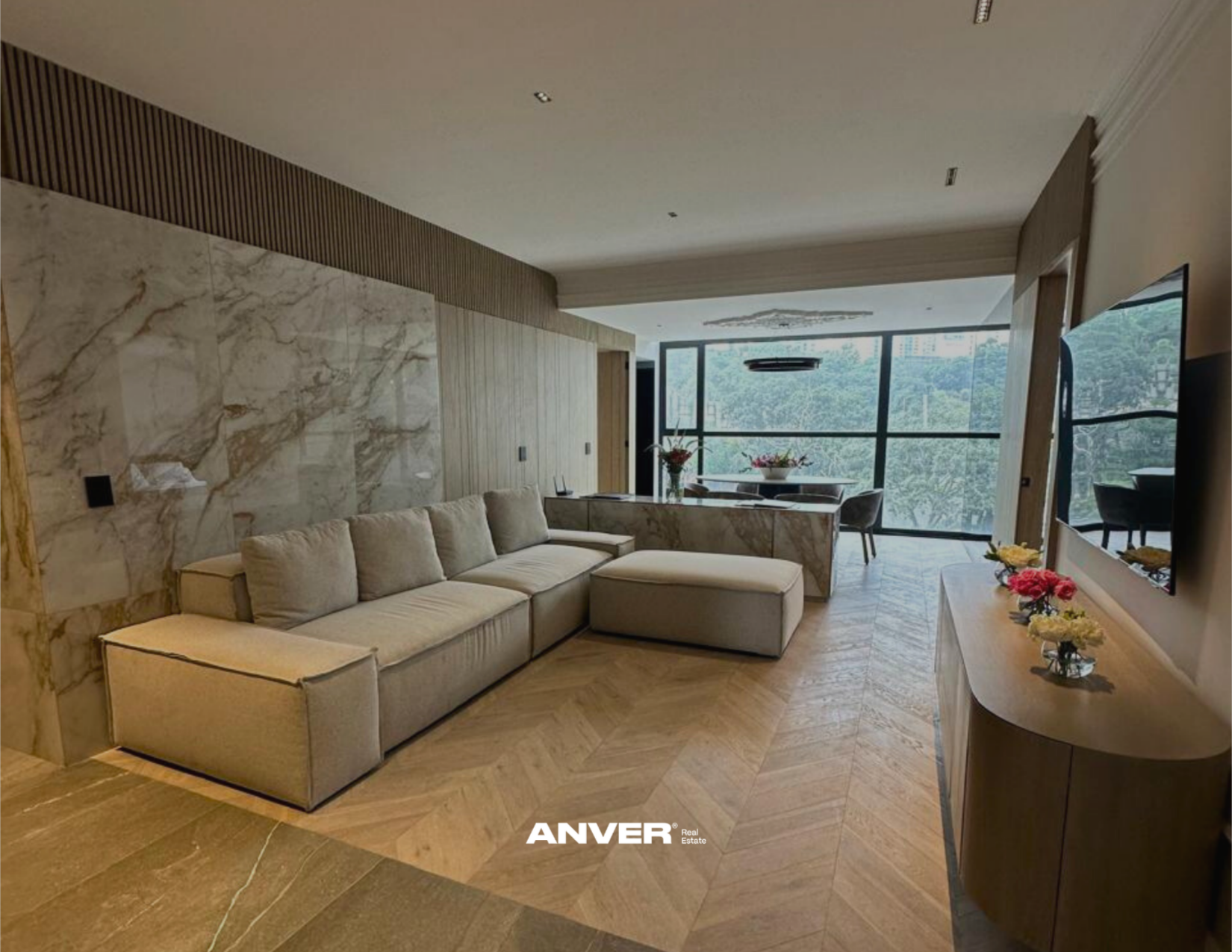 Sala moderna con sofá beige, mesa de comedor con flores, ventanas grandes con vista a árboles, decoración elegante en tonos neutros.