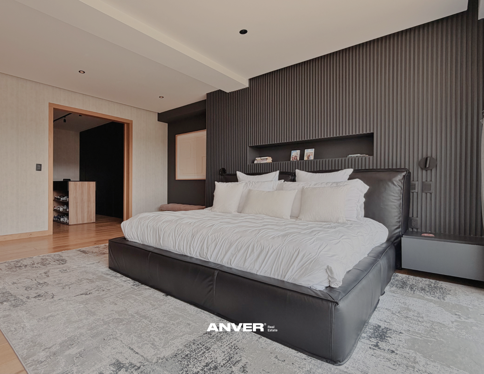 Habitación moderna con cama grande, colcha blanca y varias almohadas, paredes en tonos oscuros y muebles de madera. Alfombra gris y puerta de madera en fondo.