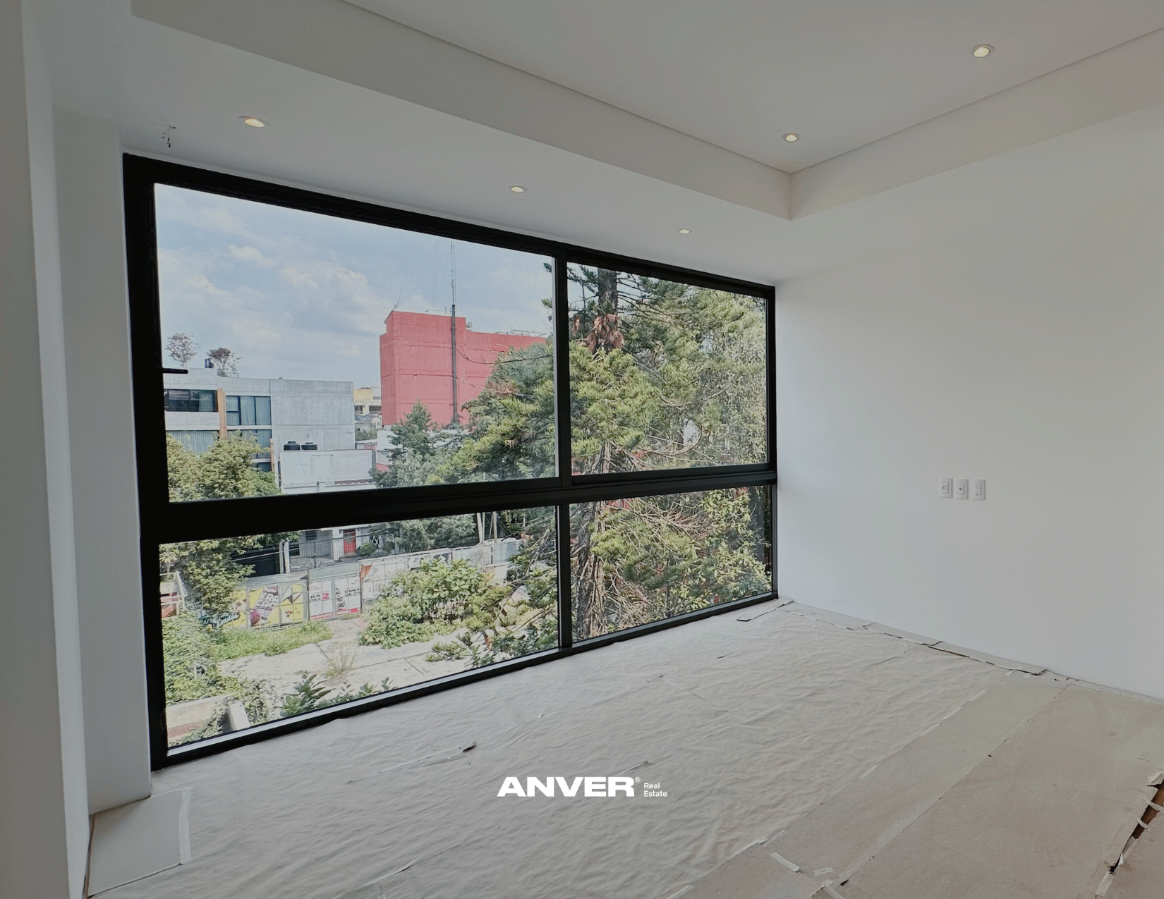 Habitación en construcción con gran ventana de piso a techo que deja ver árboles y edificios en la ciudad