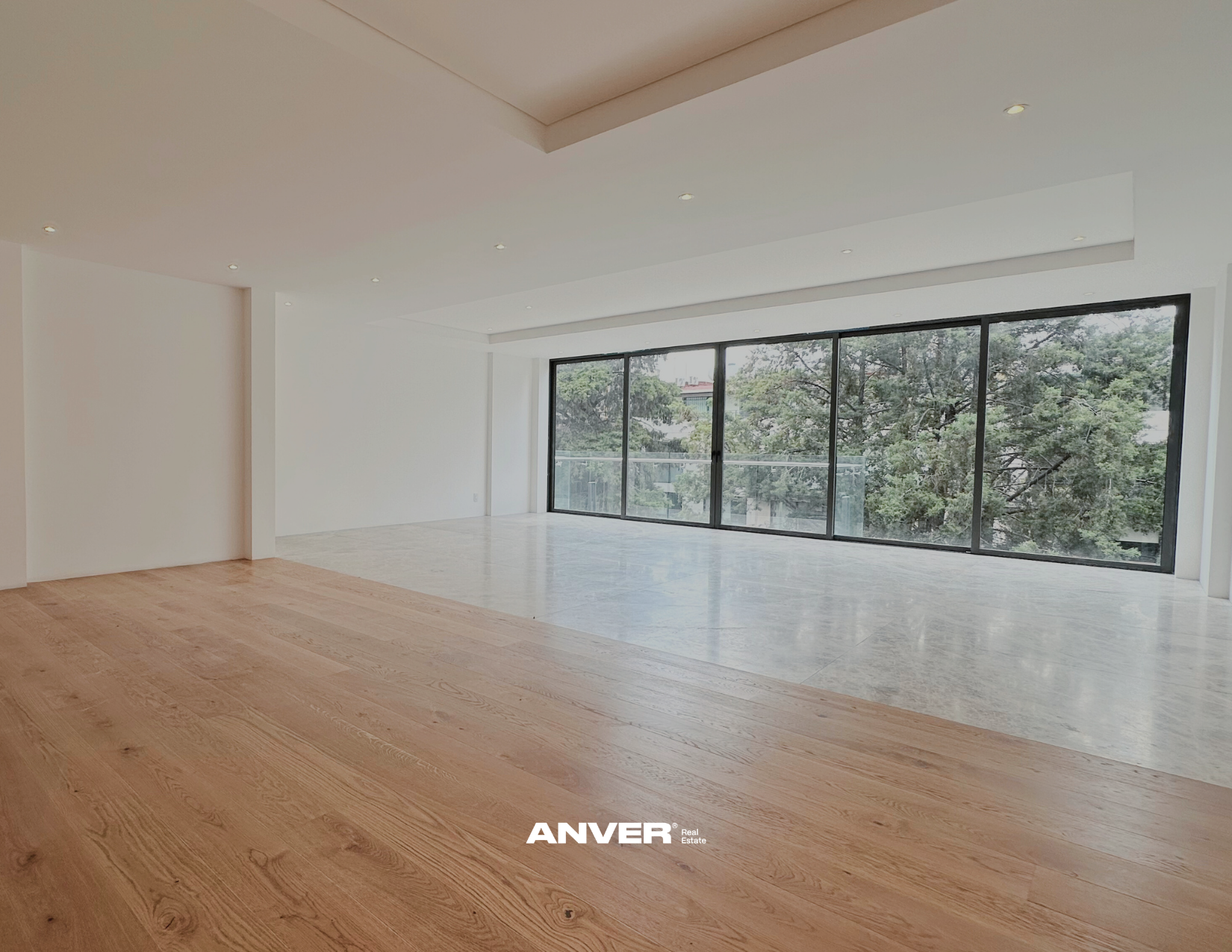 Espacio amplio y vacío con paredes blancas, piso de madera y grandes ventanales con vista a árboles.