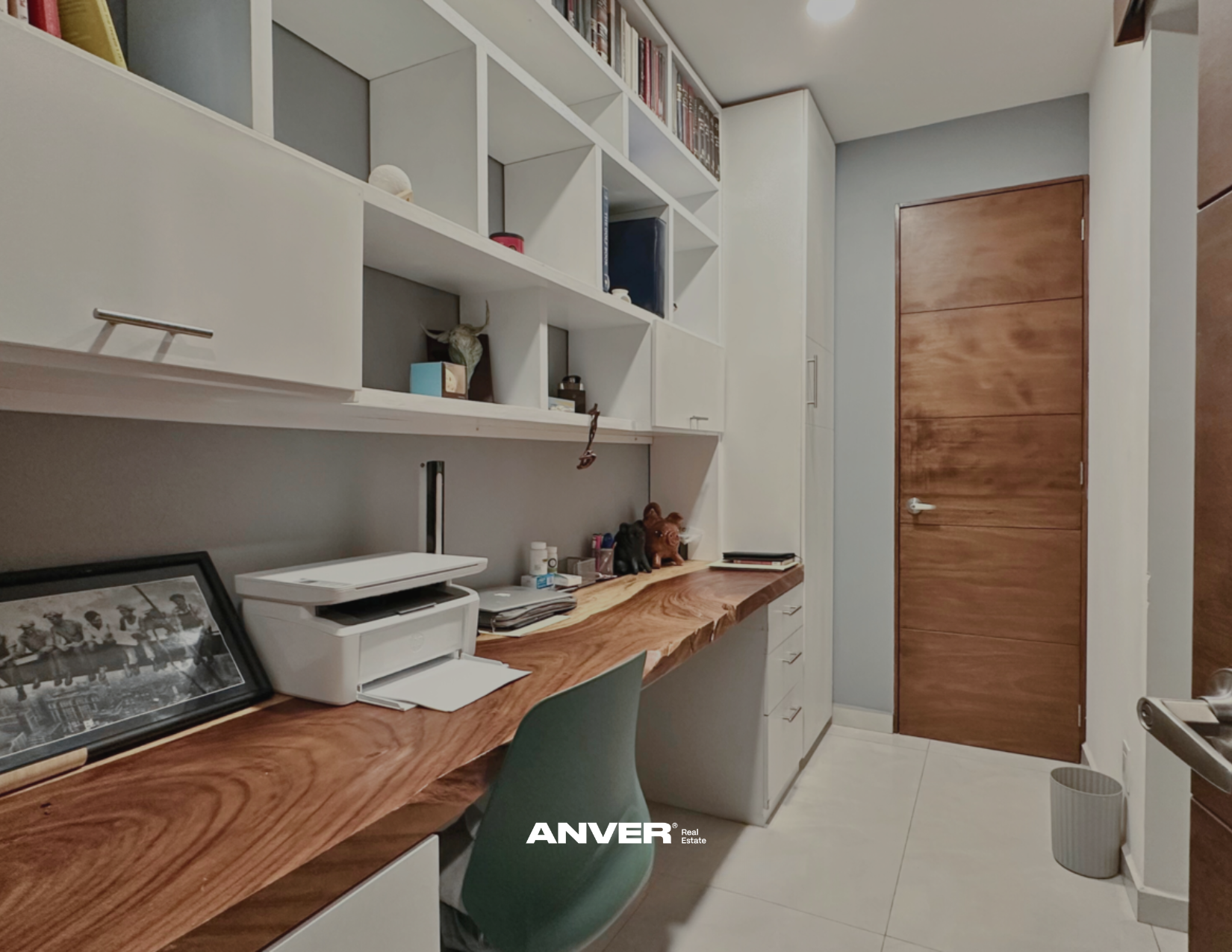 Un espacio de oficina con un escritorio de madera curva, una silla verde y estanterías blancas llenas de libros, objetos decorativos y papeles. En la esquina hay una impresora, un marco con una foto en blanco y negro, y un cubo de basura de malla metálica. La puerta es de madera y la habitación tiene paredes grises y piso claro.