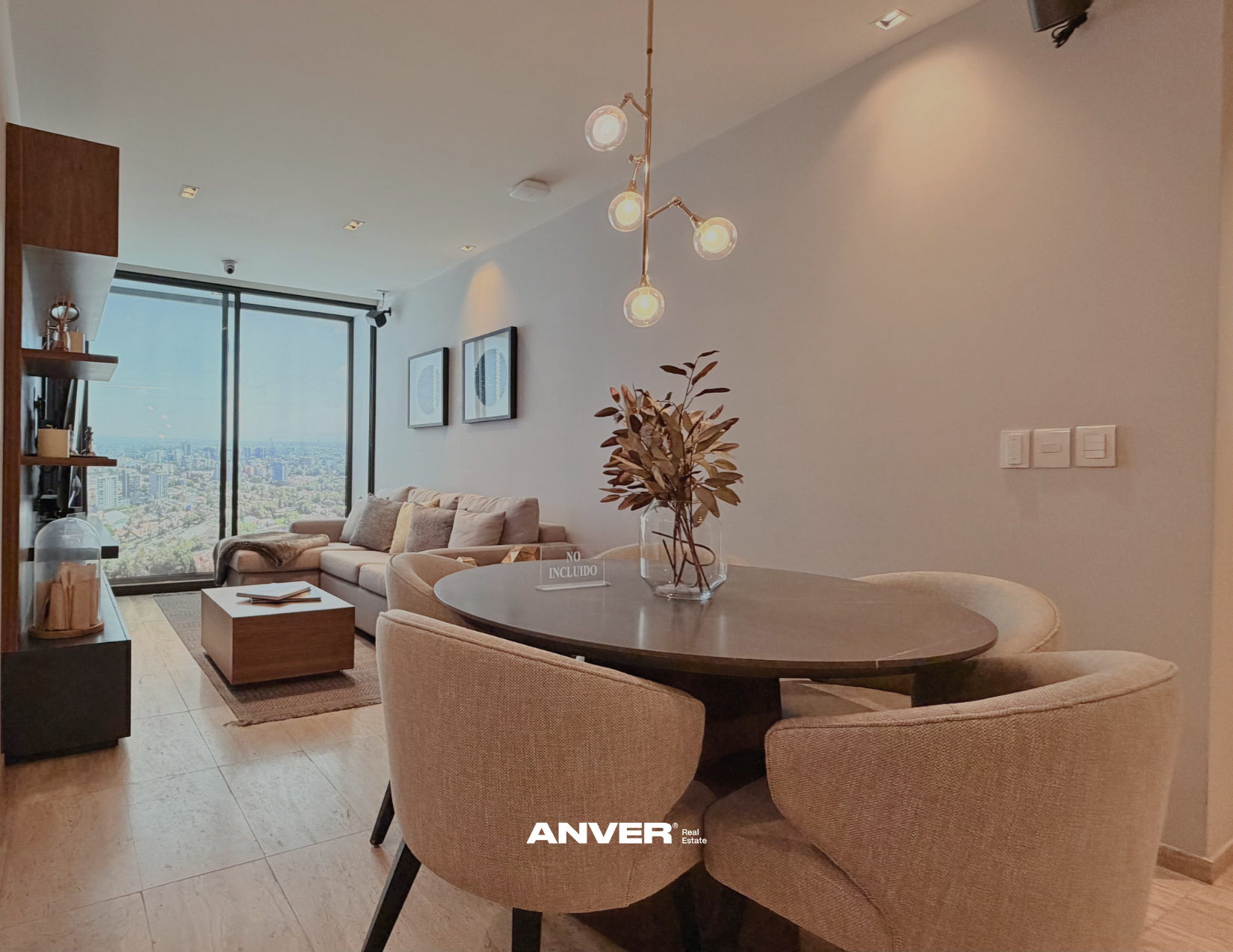 Sala moderna con vista a la ciudad, sofá beige, mesa baja de madera, lámpara de techo con bombillas expuestas, planta en jarrón de vidrio, paredes blancas con cuadros, ambiente luminoso y acogedor.