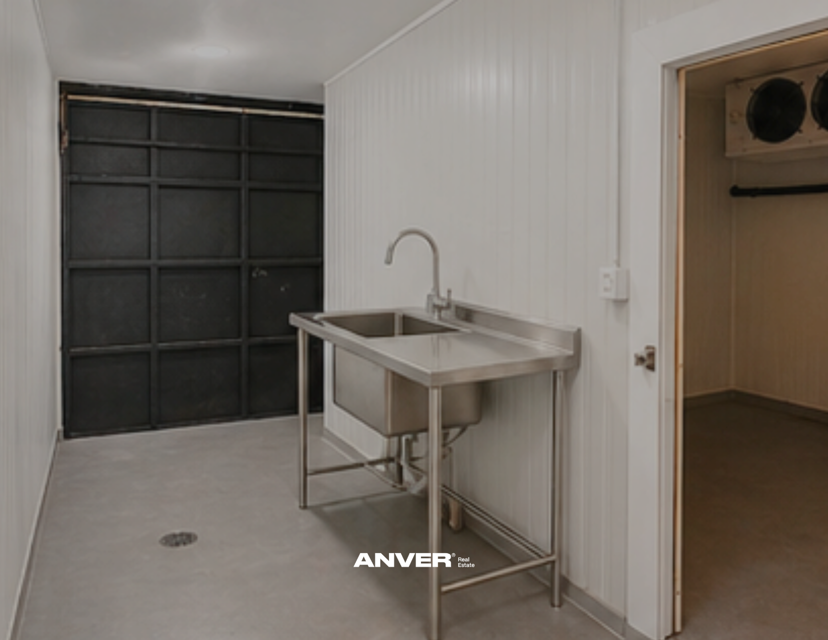 Una pequeña lavandería con lavabo de acero inoxidable, paredes blancas de paneles, puerta de garaje negra y un armario para colgar ropa.