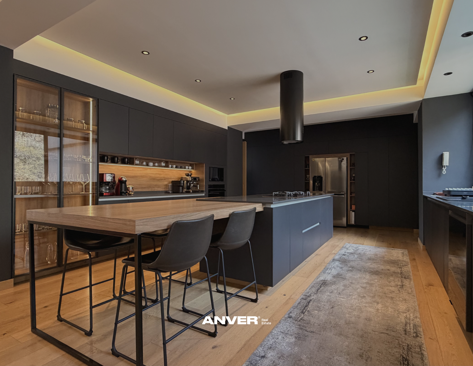 Cocina moderna con isla central, armarios oscuros, comedor con sillas negras, lámpara de extracción negra, pared gris oscuro, suelo de madera, y estufa de gas.
