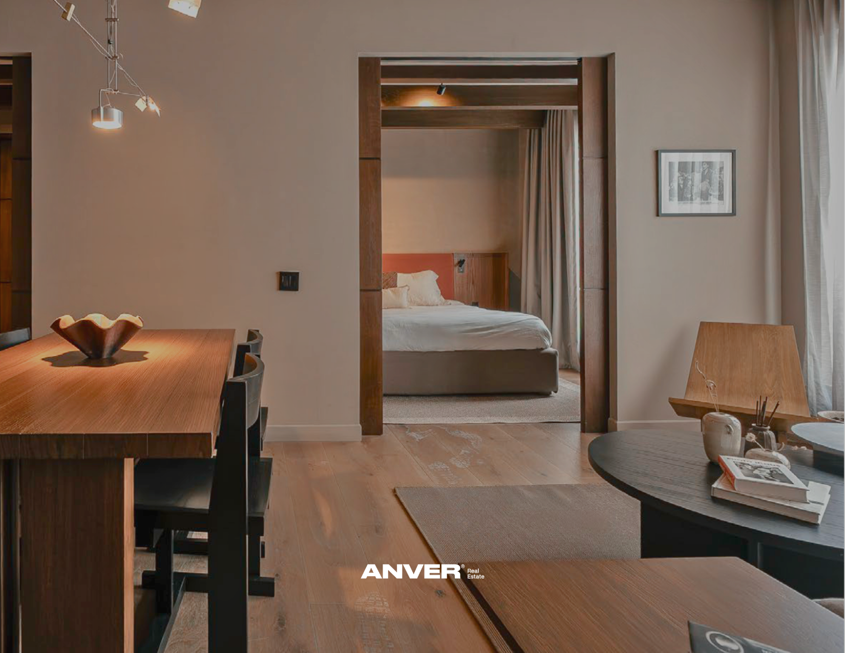 Interior de una sala y habitación con mobiliario de madera, incluyendo una mesa de comedor, sillas negras, una mesa de centro y una cama doble con cabecera de madera, cortinas beige y cuadros en la pared.