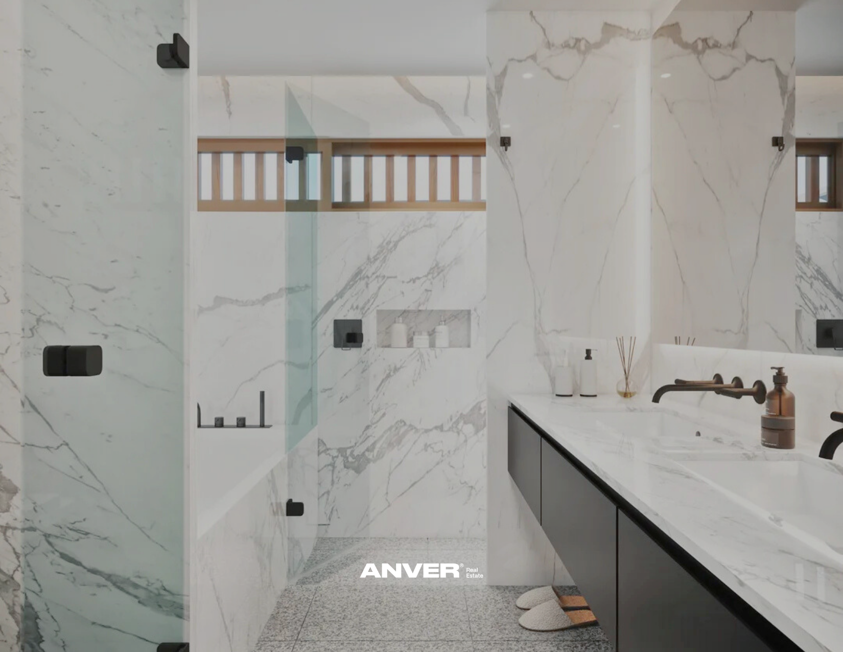 Un baño moderno con paredes y suelo de mármol blanco con vetas grises, con un lavabo sobre mueble negro, accesorios en tonos oscuros, y una ducha con una pared de cristal y accesorios en negro.
