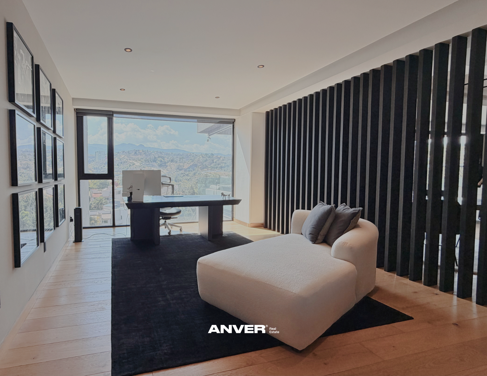 Sala con sillón beige, cojines gris, escritorio con computadora y vista panorámica de ciudad y montañas.