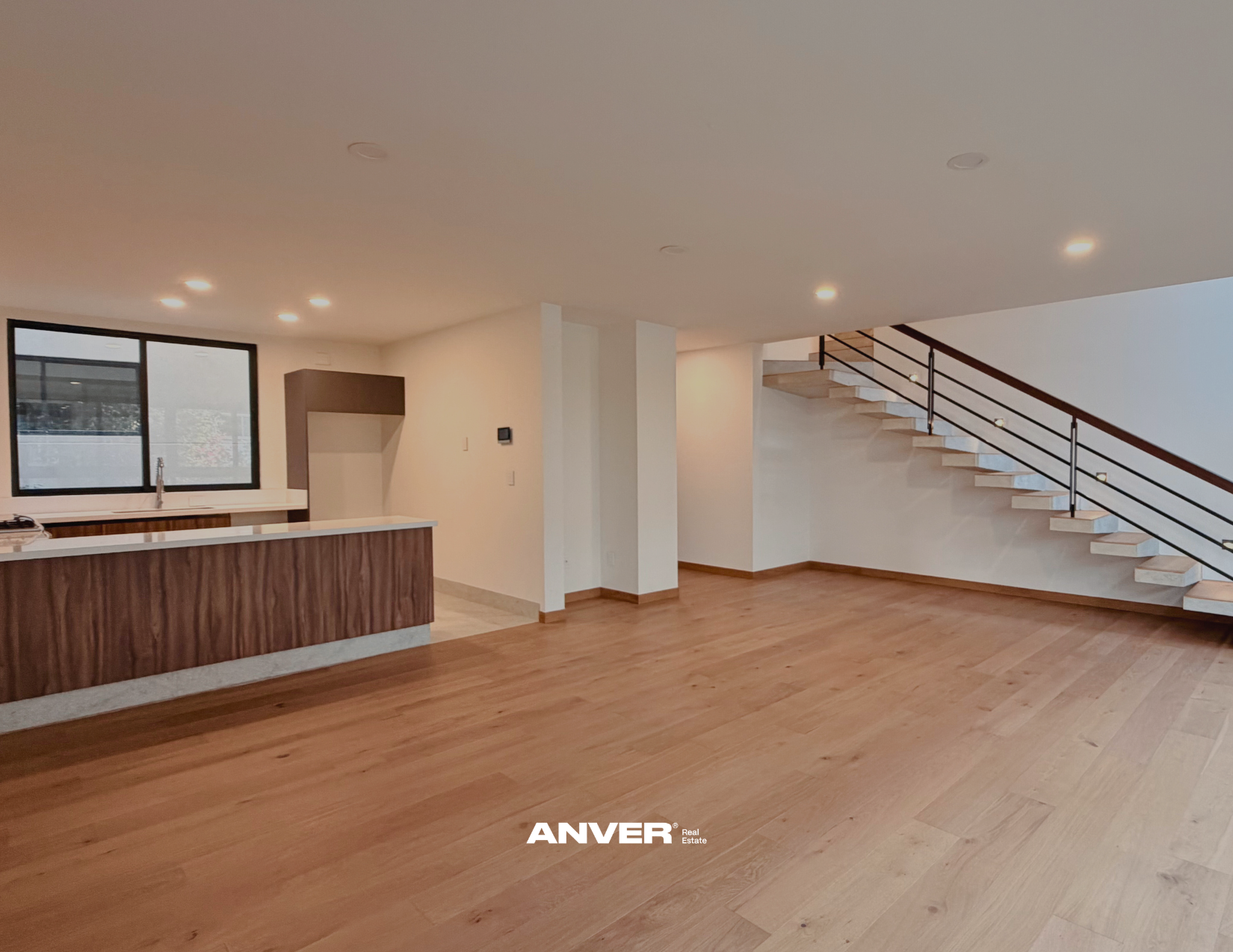 Interior de una casa moderna con sala y cocina, escaleras de madera, paredes blancas, pisos de madera clara, ventanas grandes y luces empotradas en el techo.