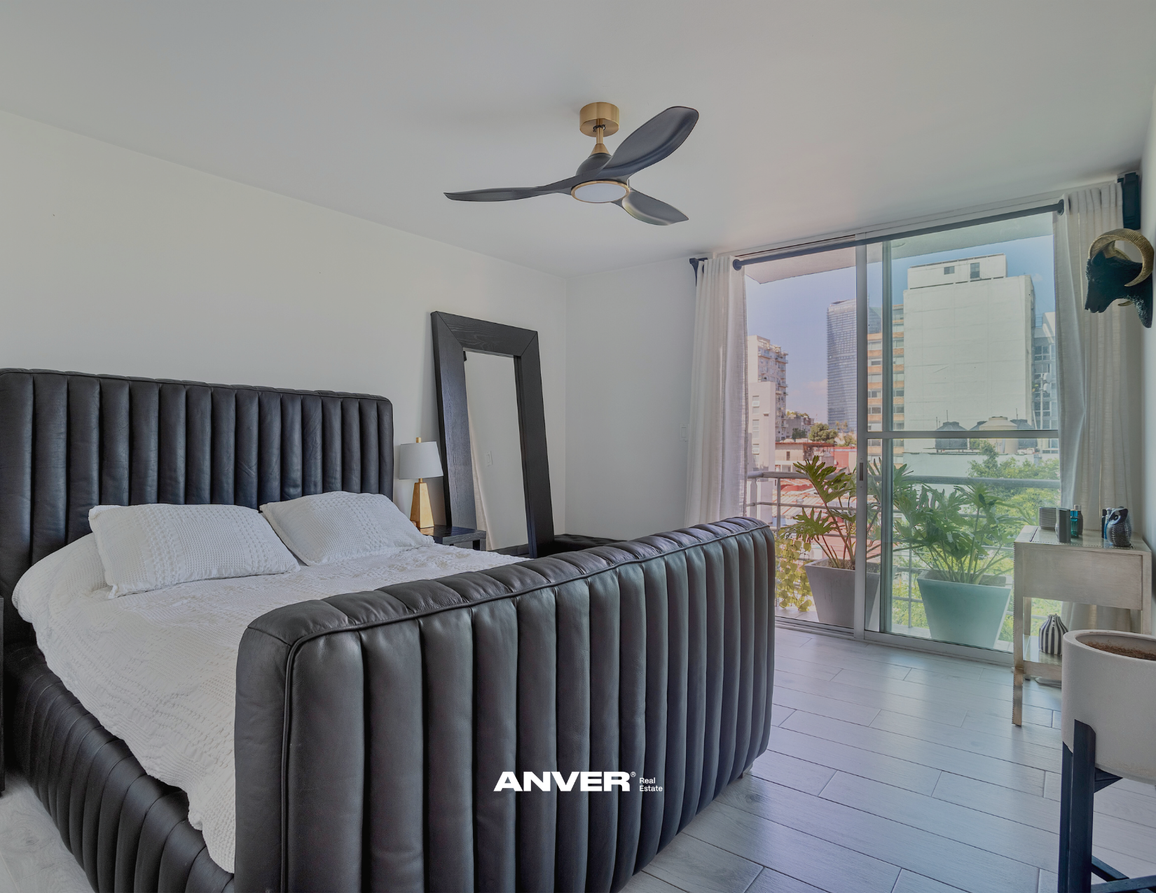 Habitación moderna con cama con cabecera de cuero, espejo de marco negro, lámpara de mesa, plantas en balcón y vista urbana desde la ventana.