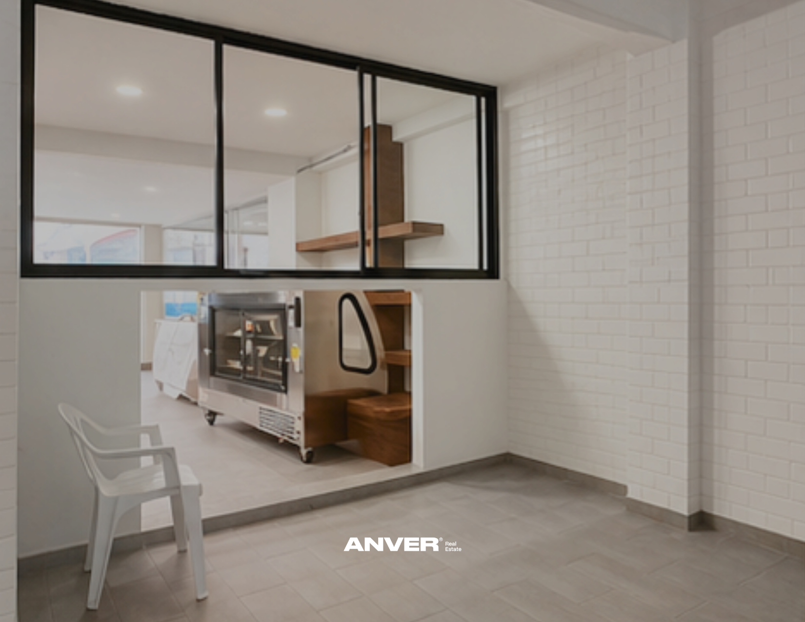 Interiores de un espacio moderno con paredes blancas y una ventana de cristal con marco negro, que permite ver otra área. Incluye sillas blancas, un mobiliario de cocina con acero inoxidable y detalles en madera, y un piso de azulejos gris claros.