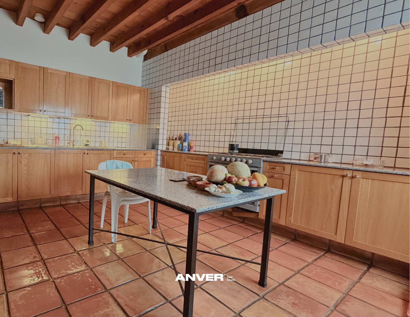 Cocina con gabinetes de madera, baldosas en las paredes y piso de terracota, mesa en el centro con frutas y verduras.