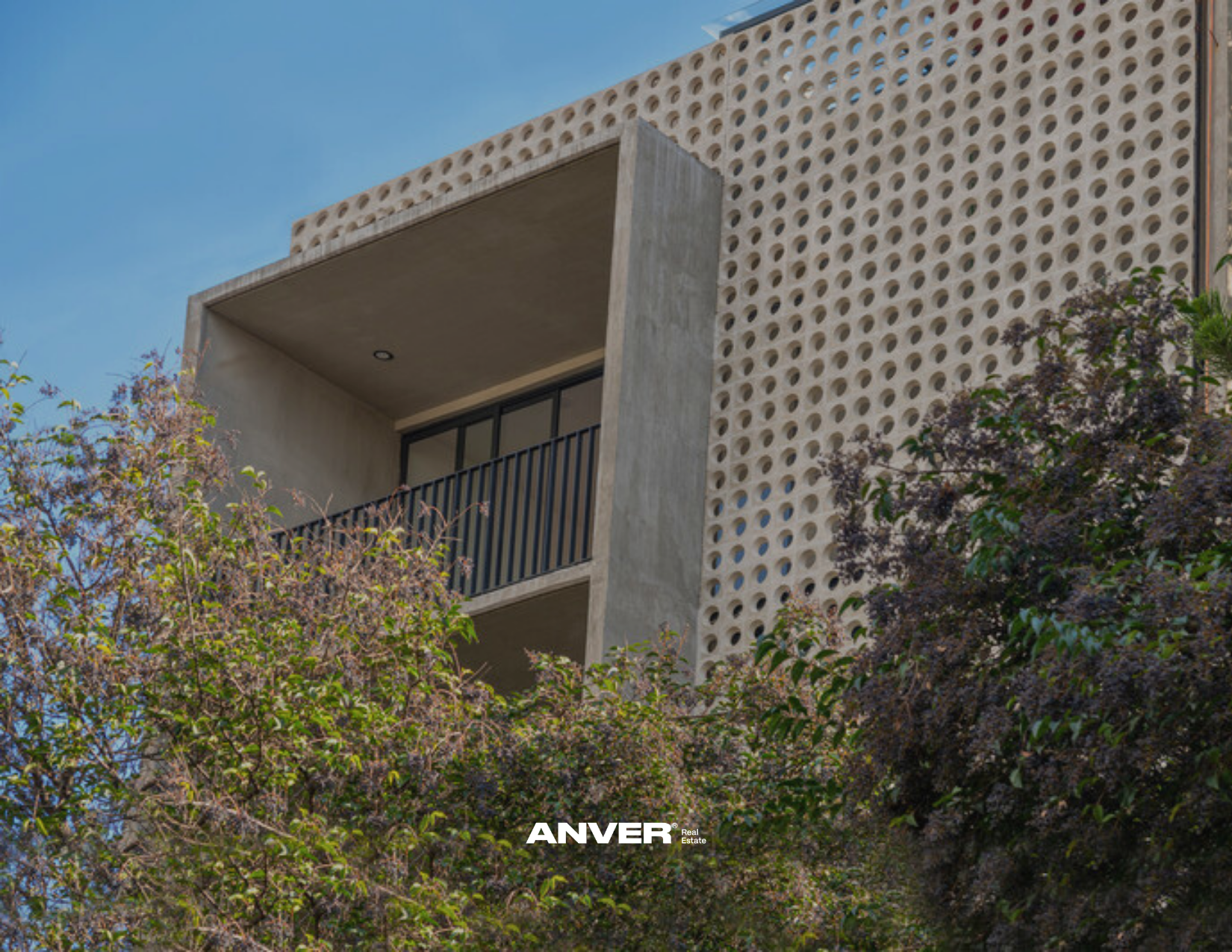Edificio moderno con balcón y fachada de bloques de concreto con agujeros circulares, rodeado de árboles con follaje denso, bajo un cielo despejado.