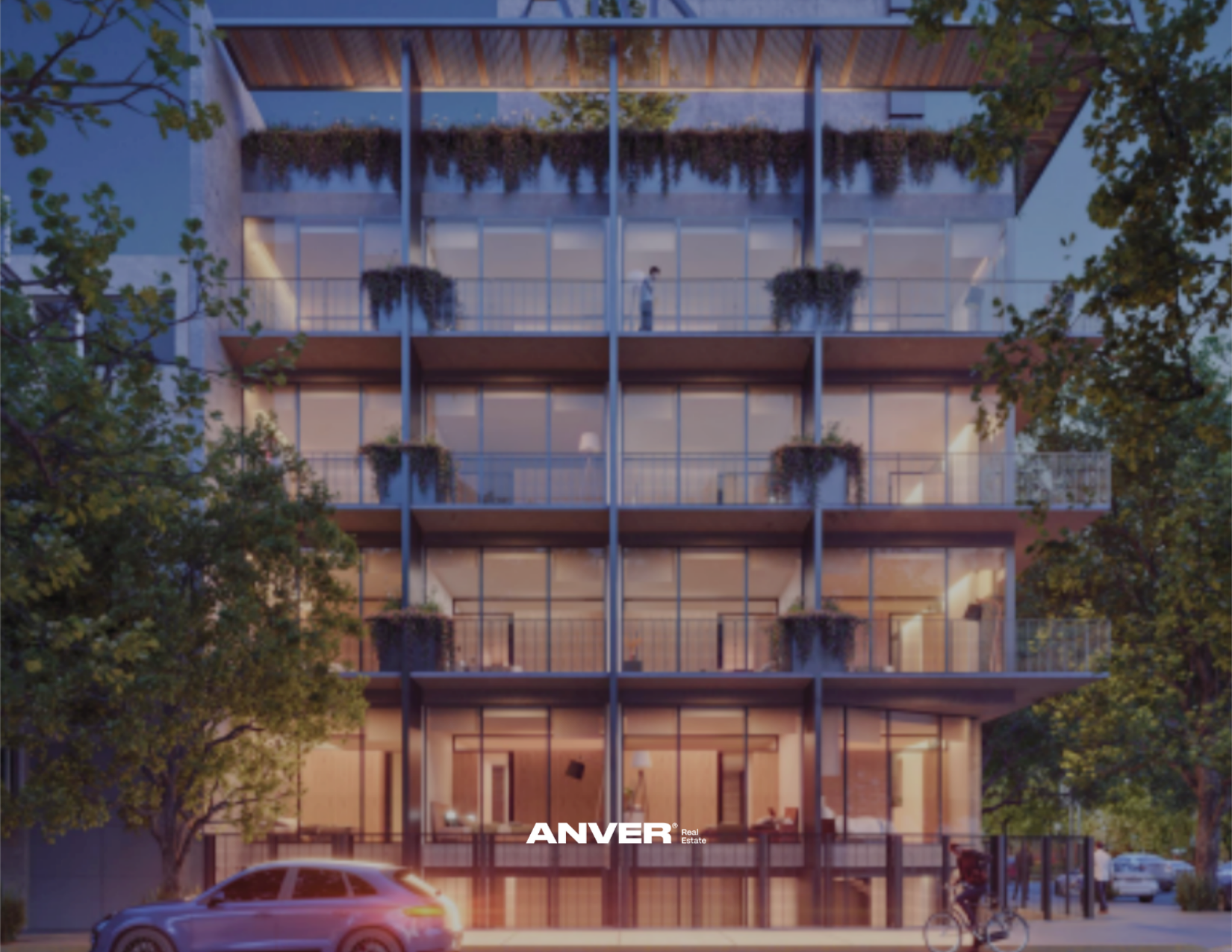 Edificio moderno de varios pisos con balcones llenos de plantas y grandes ventanas de cristal, visto en la noche con luces interiores encendidas, rodeado de árboles y con autos y bicicleta en la calle del frente.