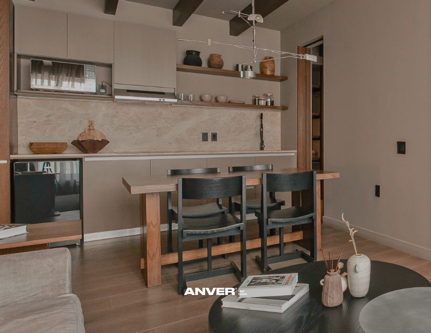 Interior de una sala o comedor con mesa de madera y sillas negras, y una pared de cocina con estantes y utensilios de cocina decorativos. Hay una mesa de centro con libros y decoraciones de cerámica.