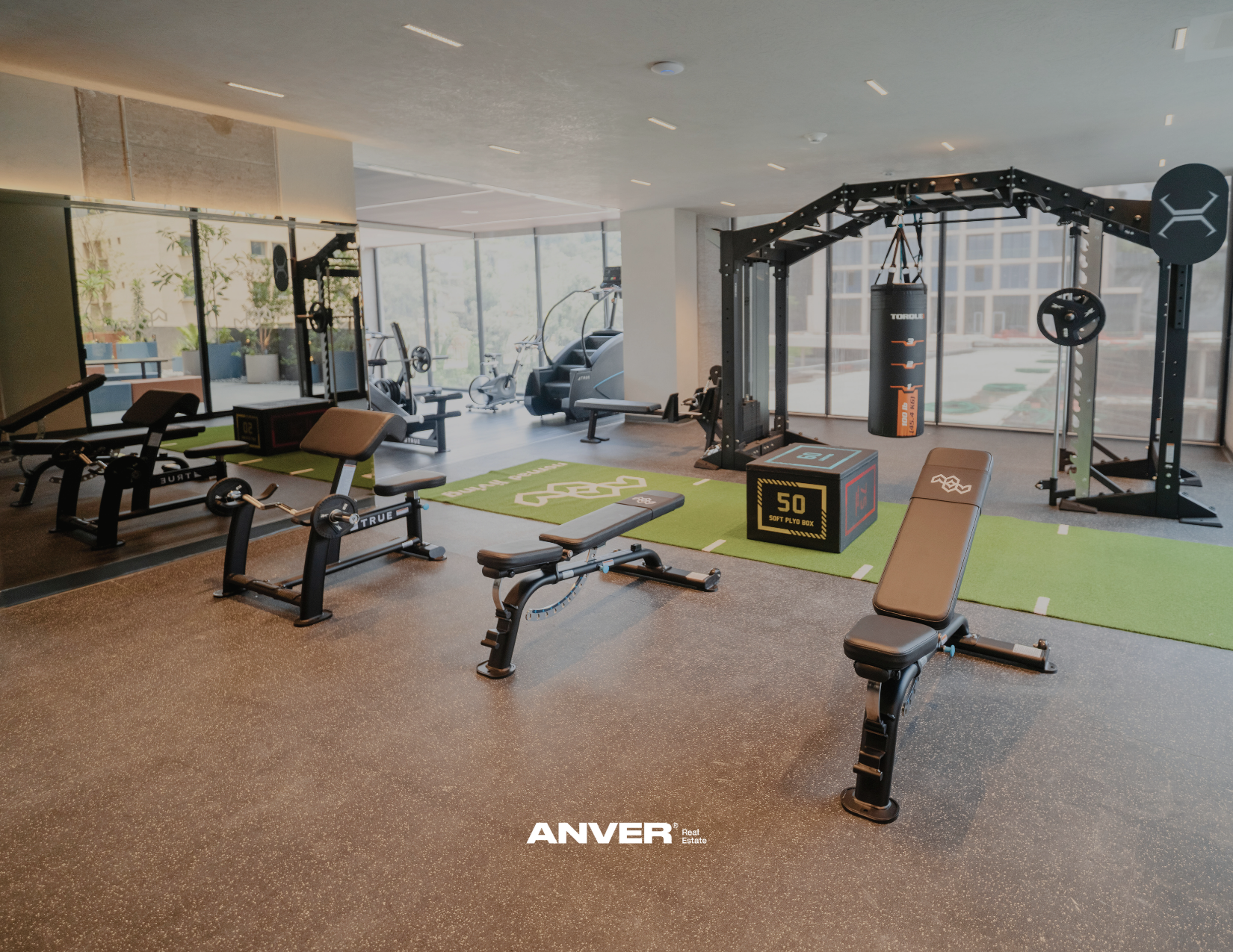 Gimnasio con máquinas de pesas, colchonetas verdes y saco de boxeo en una habitación moderna con ventanas grandes y vista a la ciudad.